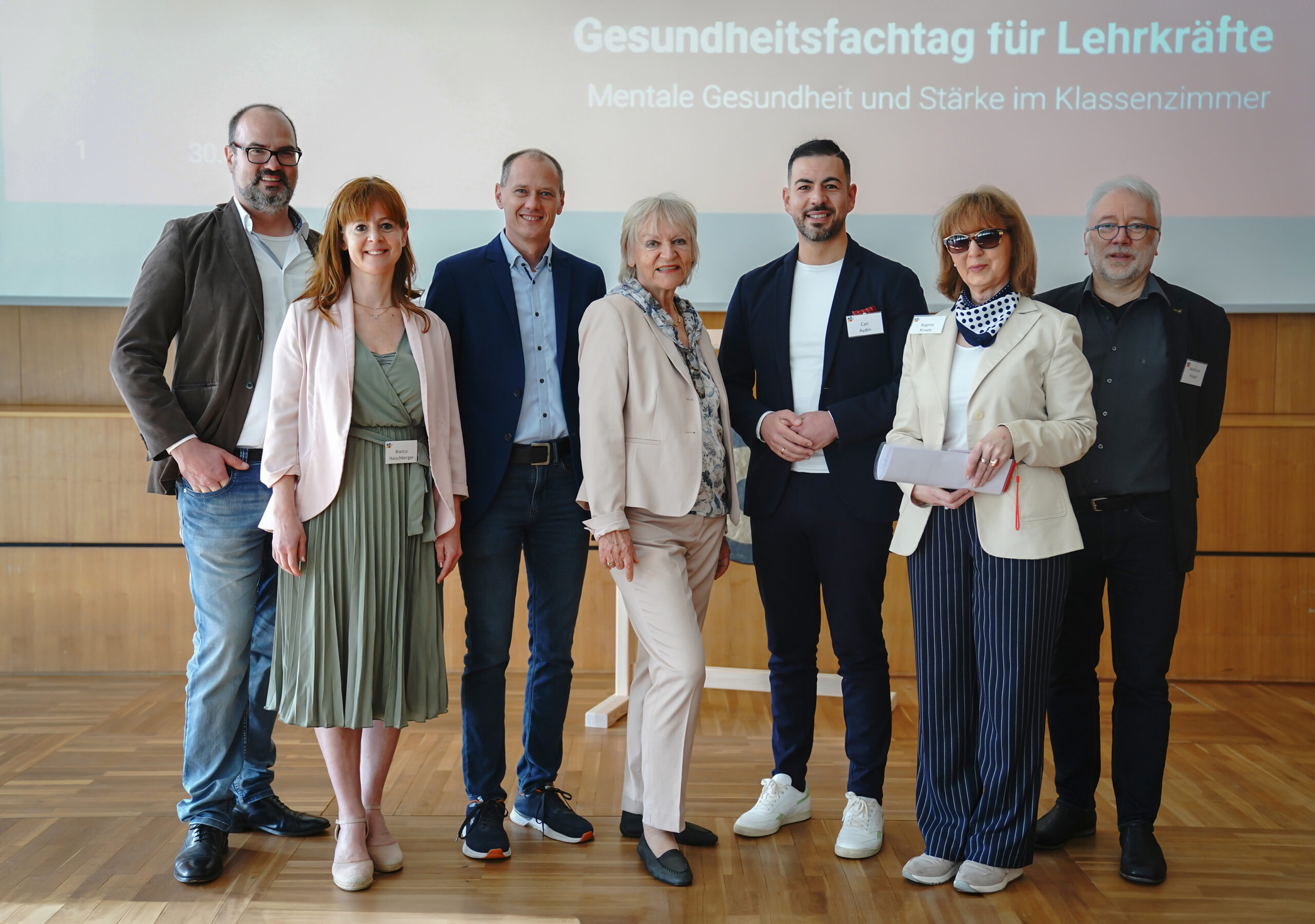 A group of seven people stands in front of a screen that reads "Gesundheitsfachtag für Lehrkräfte" in German, representing Hochschule Coburg. They are dressed in smart casual attire, smiling, and posing indoors on wooden flooring.