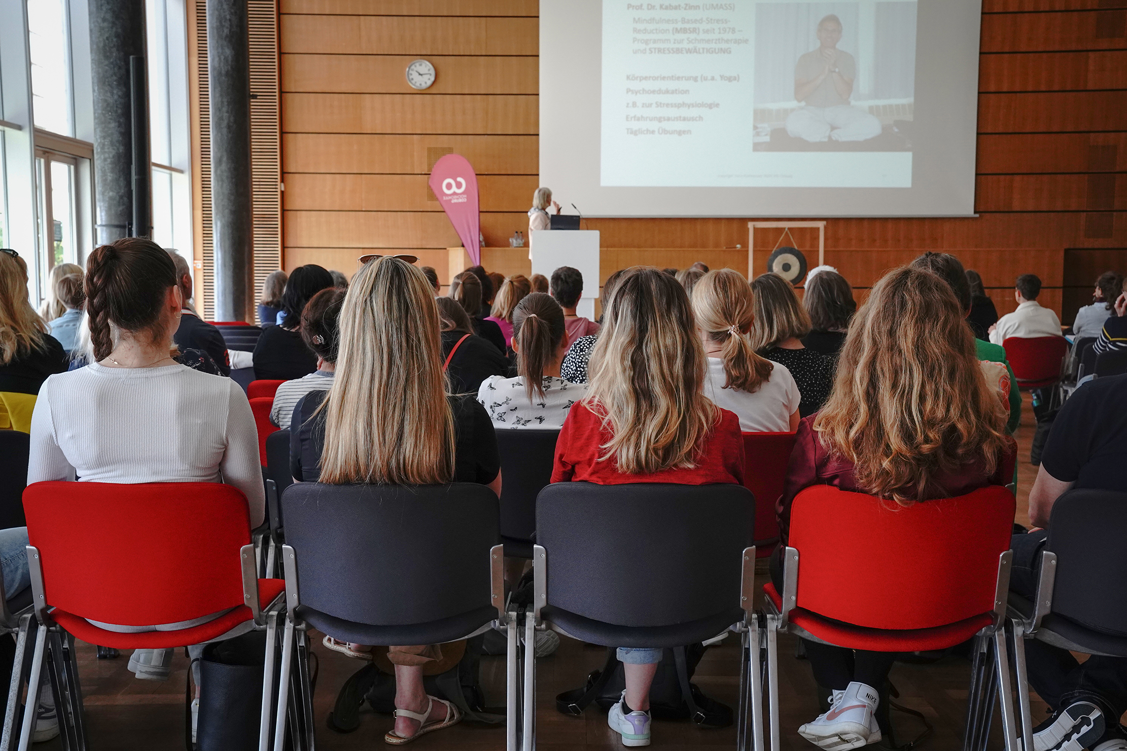 Audience sitting on chairs, facing a speaker at a podium in a conference room at Hochschule Coburg. A presentation is projected on a screen, with the university banner displayed to the side.