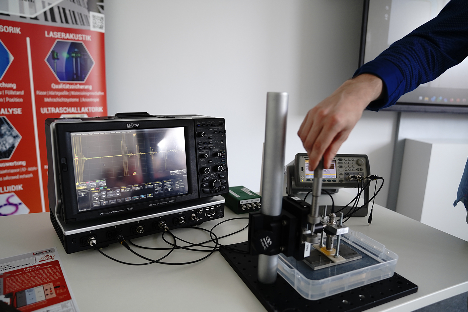 A person is expertly operating an oscilloscope and other electronic testing equipment on a table, surrounded by wires and a probe. A screen reveals detailed waveform data. In the background, red promotional banners proudly display Hochschule Coburg's emblem, adding an academic touch to the scene.