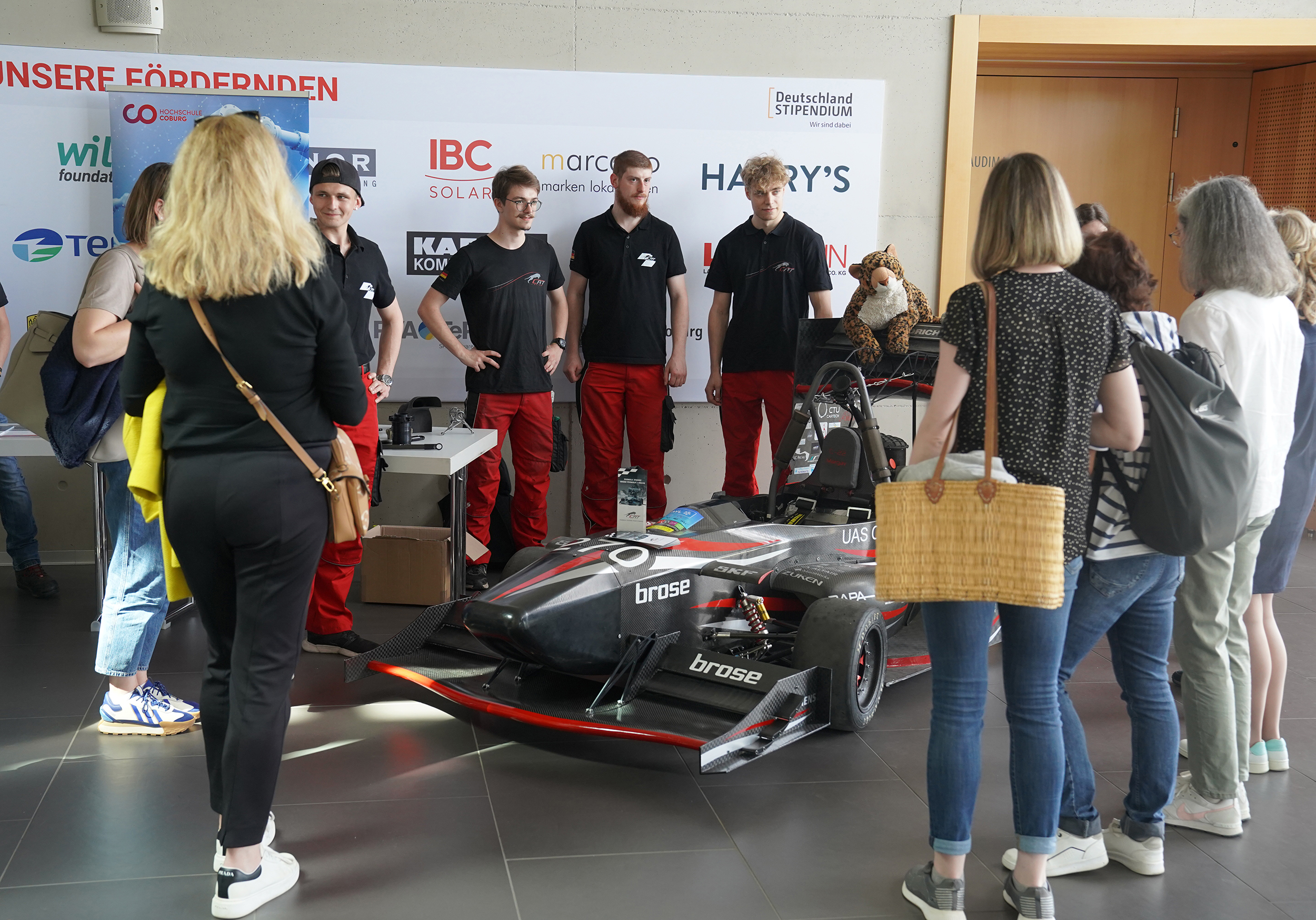 A group of people stands around a black and red race car displayed indoors. Four individuals in black shirts and red pants from Hochschule Coburg are presenting the car. Banners and sponsor logos, including Hochschule Coburg's emblem, are visible on the wall behind them.