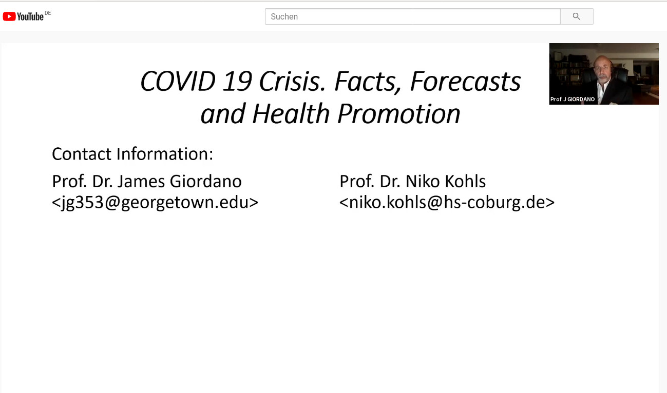 Auf der Folie mit dem Titel „COVID-19-Krise: Fakten, Prognosen und Gesundheitsförderung“ sind die Kontaktdaten von Prof. Dr. James Giordano und Prof. Dr. Niko Kohls, einschließlich ihrer E-Mail-Adressen, zu sehen. In der oberen rechten Ecke erscheint ein Videoanruffenster mit einem Mann, der möglicherweise von der Hochschule Coburg sendet.