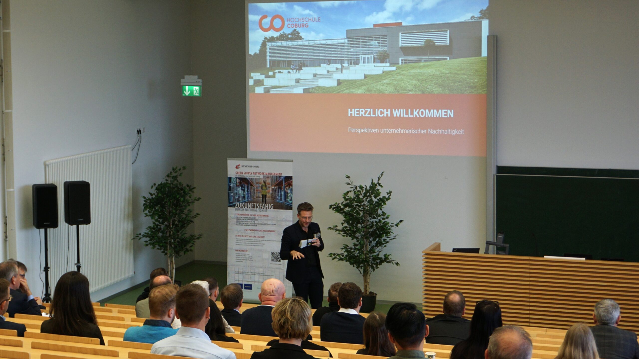 A speaker presents to an audience in a lecture hall at Hochschule Coburg. A large screen displays a building image and the text "Herzlich Willkommen." Attendees are seated on wooden benches, with plants flanking the stage.