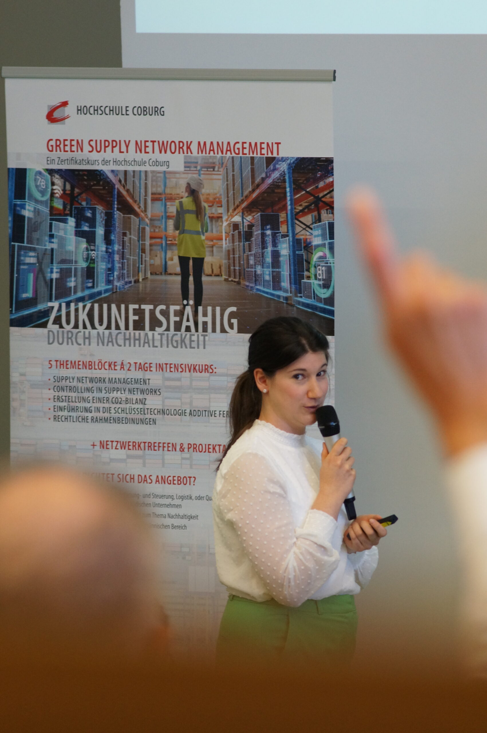 A presenter at Hochschule Coburg holds a microphone, standing before a "Green Supply Network Management" poster. Digital warehouse graphics adorn the backdrop, while an audience member's arm is raised inquisitively in the foreground.