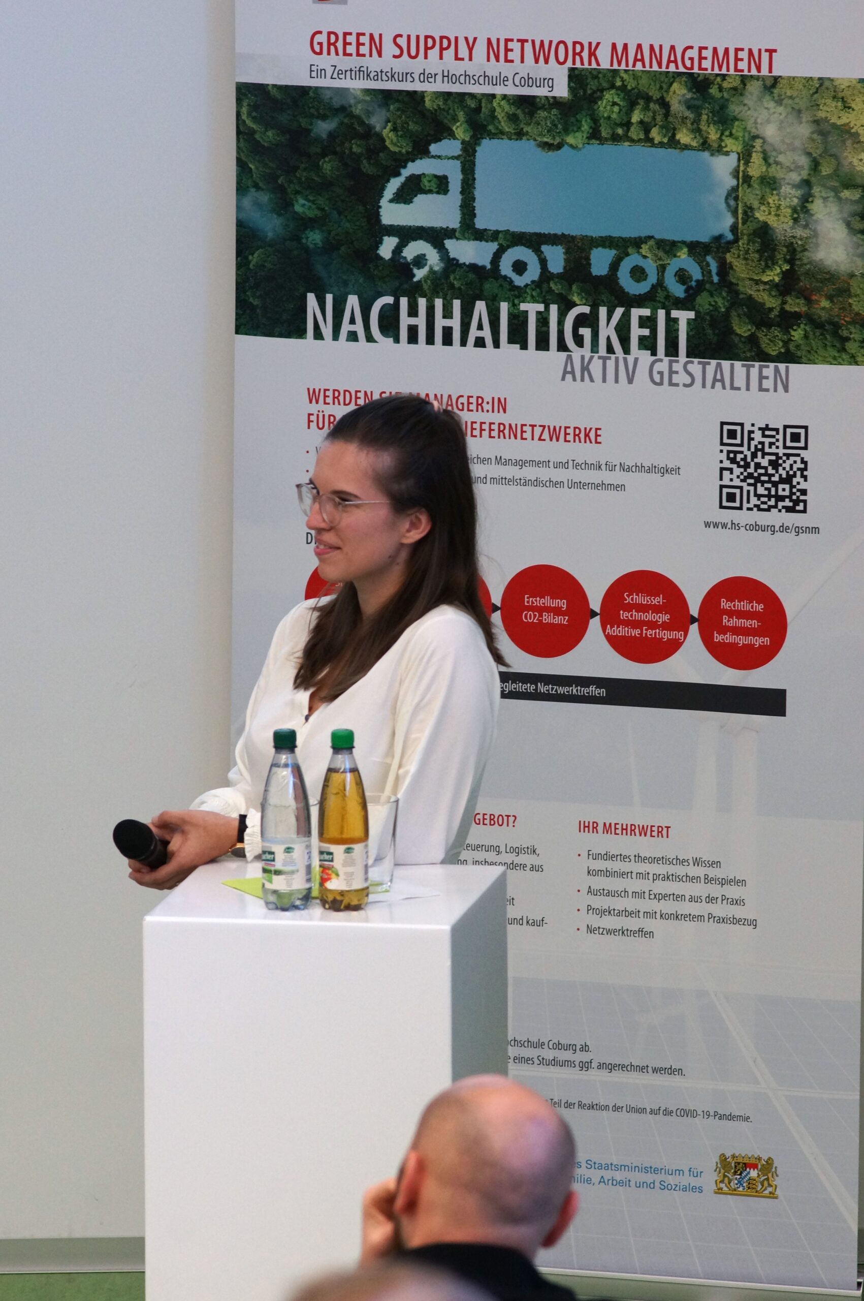 A woman stands in front of a Hochschule Coburg poster about sustainability management at an event. She holds a microphone and a notebook, with two bottles on a small table beside her. The poster, featuring a truck icon and text in German, underscores the university's commitment to sustainable practices.