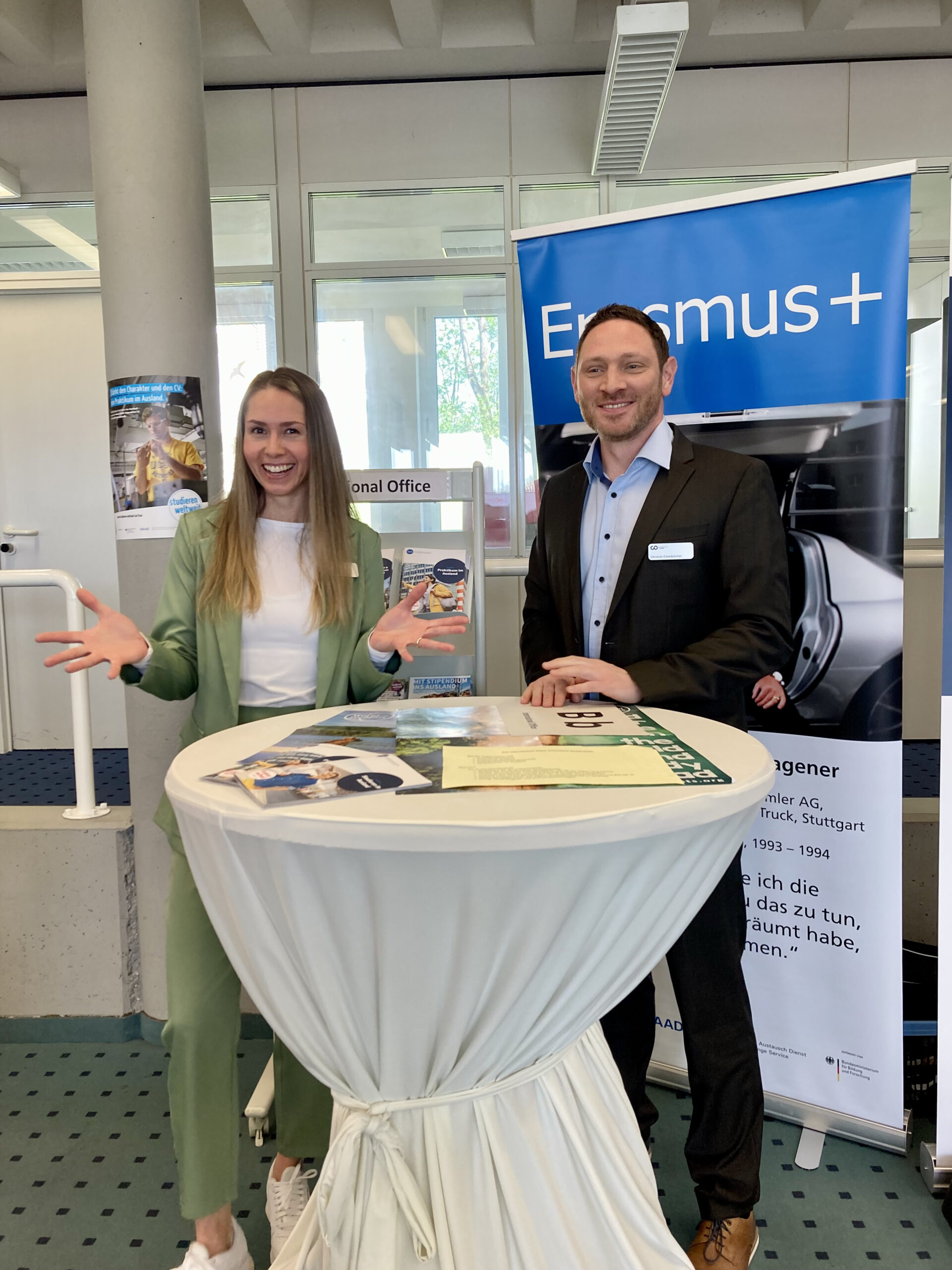 Zwei Personen stehen an einem runden Tisch am Erasmus+ Stand der Hochschule Coburg. Die Frau gestikuliert mit den Händen, der Mann lächelt. Im Hintergrund ist ein blaues Erasmus+ Banner zu sehen, auf dem Tisch liegt eine Broschüre zum Thema Auslandsstudium.