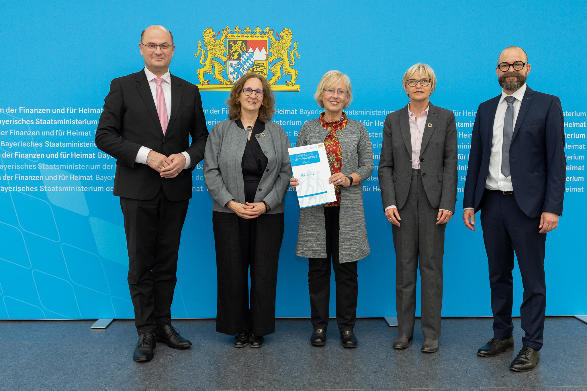 Fünf Personen in formeller Kleidung stehen vor einem blauen Hintergrund mit Text der Hochschule Coburg. Eine Person in der Mitte hält mit beiden Händen ein Dokument, über ihnen prangt prominent das Wappen-Emblem.