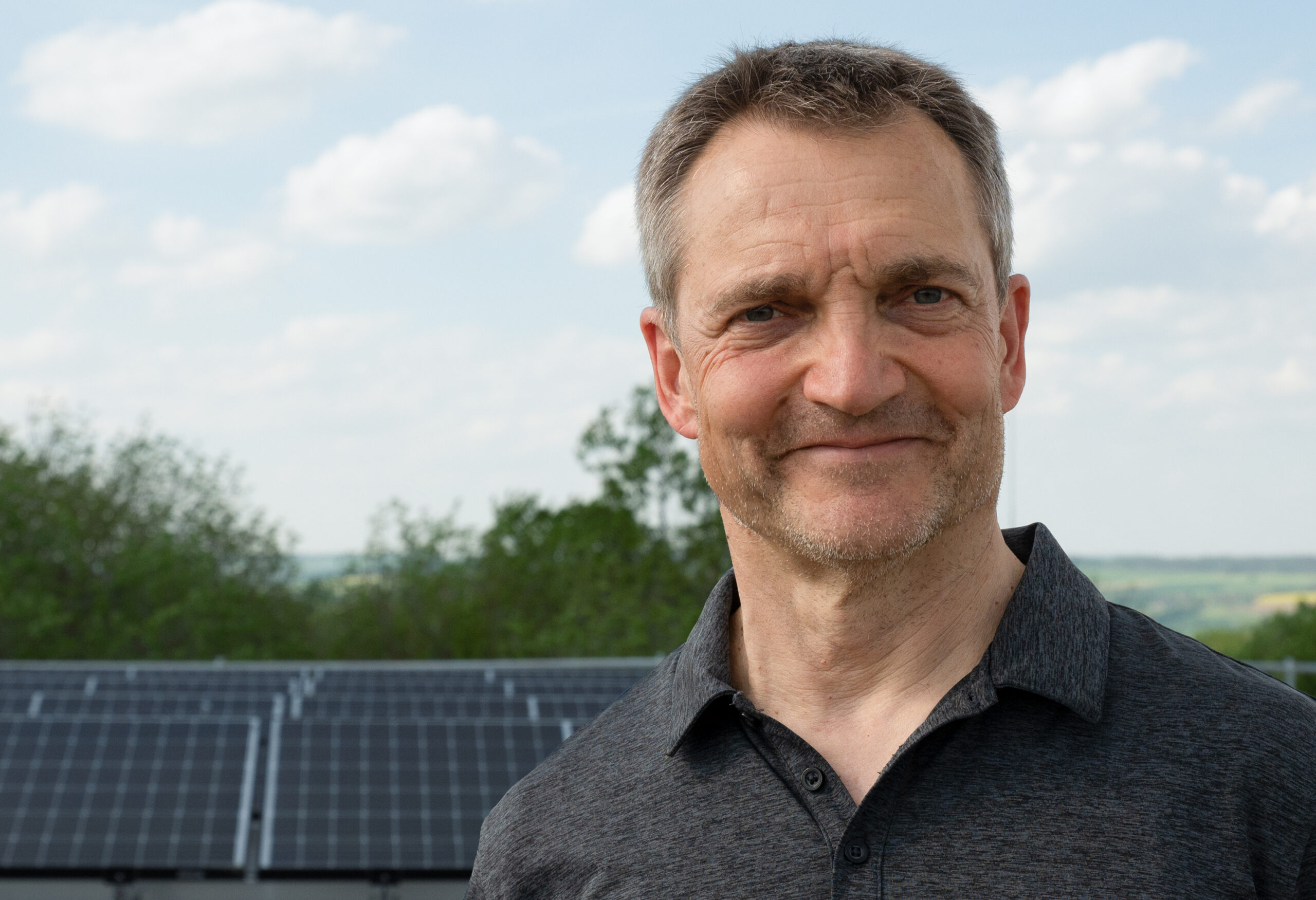 Ein Mann in einem dunklen Poloshirt steht im Freien und lächelt stolz vor den Solarmodulen auf dem Campus der Hochschule Coburg. Der Hintergrund zeigt einen teilweise bewölkten Himmel und üppiges Grün, was sowohl Innovation als auch Natur einfängt.