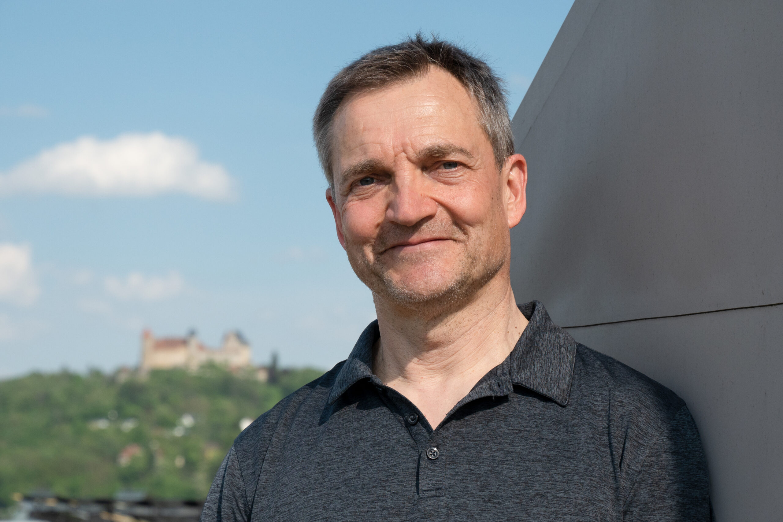 Ein Mann in einem dunklen Poloshirt steht im Freien und lehnt an einer hellen Wand. In der Ferne, hinter der grünen Landschaft, liegt die Hochschule Coburg unter einem blauen Himmel mit weißen Wolken.