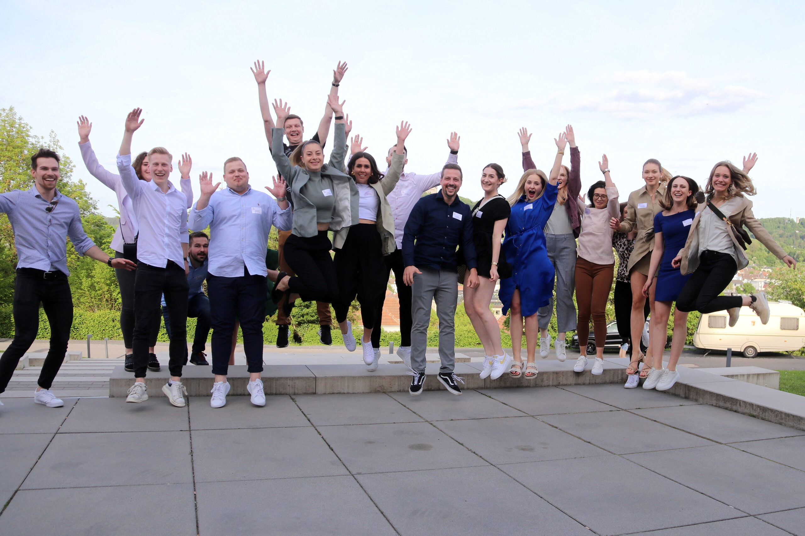 Eine Gruppe von Menschen in legerer Business-Kleidung springt im Gleichschritt auf einer gepflasterten Fläche mit malerischer Aussicht. Sie wirken fröhlich, während sie in der Nähe der Hochschule Coburg feiern. In der Ferne runden ein Wohnmobil und malerische Hügel die lebhafte Szene ab.