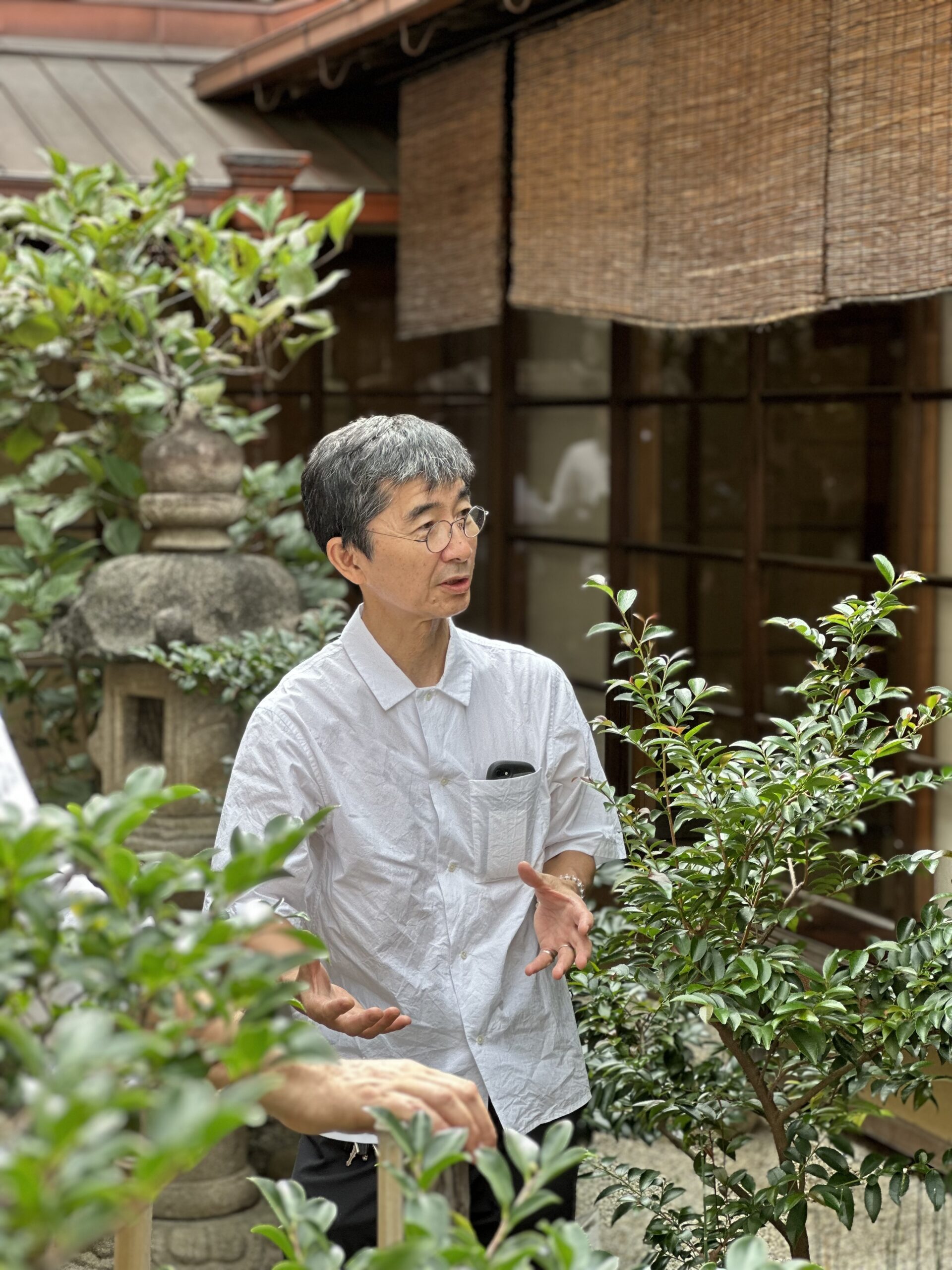 A person in a white shirt speaks animatedly in a lush garden with green plants and a stone lantern, evoking the serene ambiance of Hochschule Coburg. The backdrop features a traditional-style building with wooden windows and woven shades.