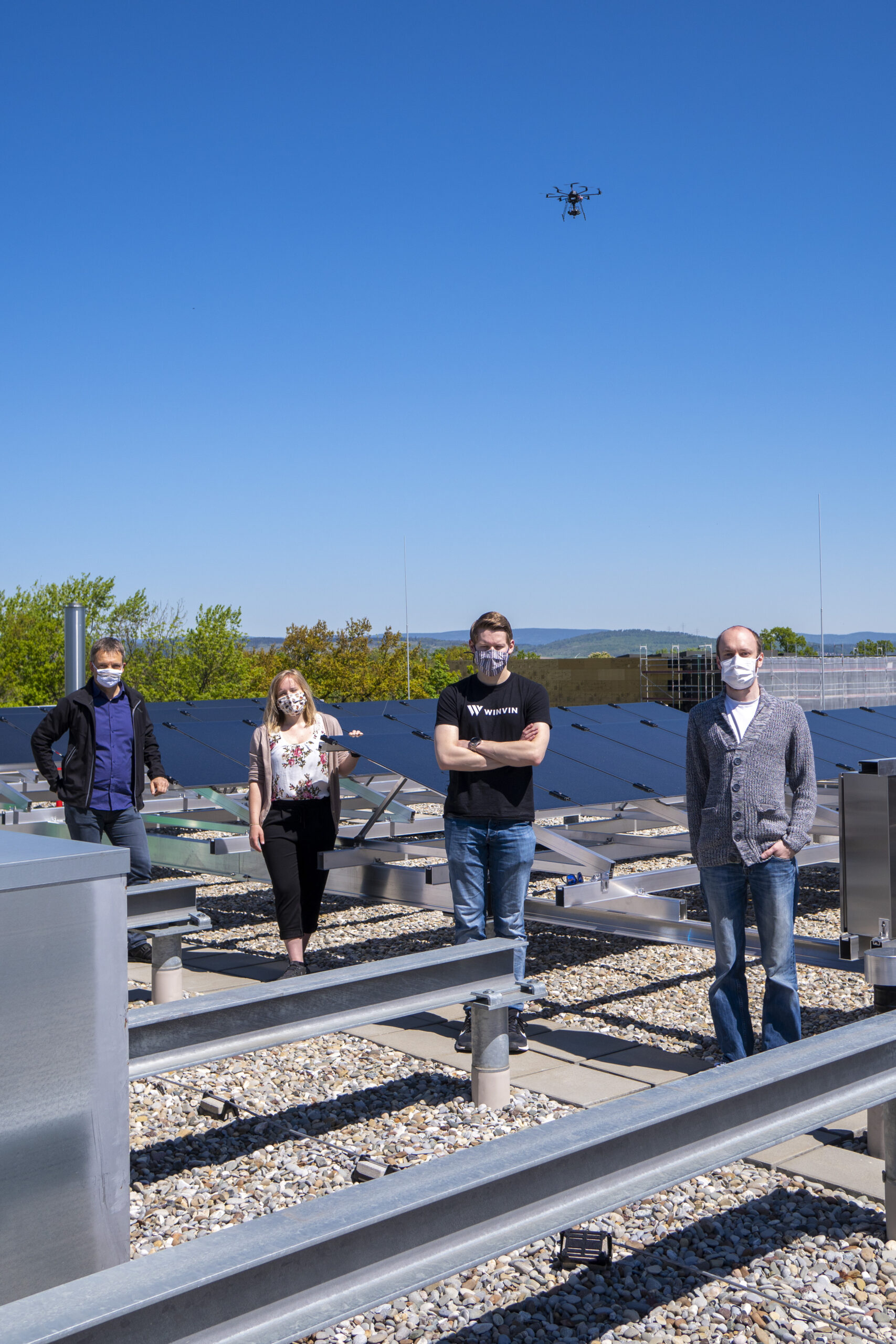 Vier Menschen mit Masken stehen auf einem Dach mit Solarmodulen der Hochschule Coburg. Über ihnen schwebt eine Drohne im klaren blauen Himmel. Lässig gekleidet genießen sie den sonnigen Tag mit viel Grün im Hintergrund und verbinden so Technologie und Natur nahtlos miteinander.