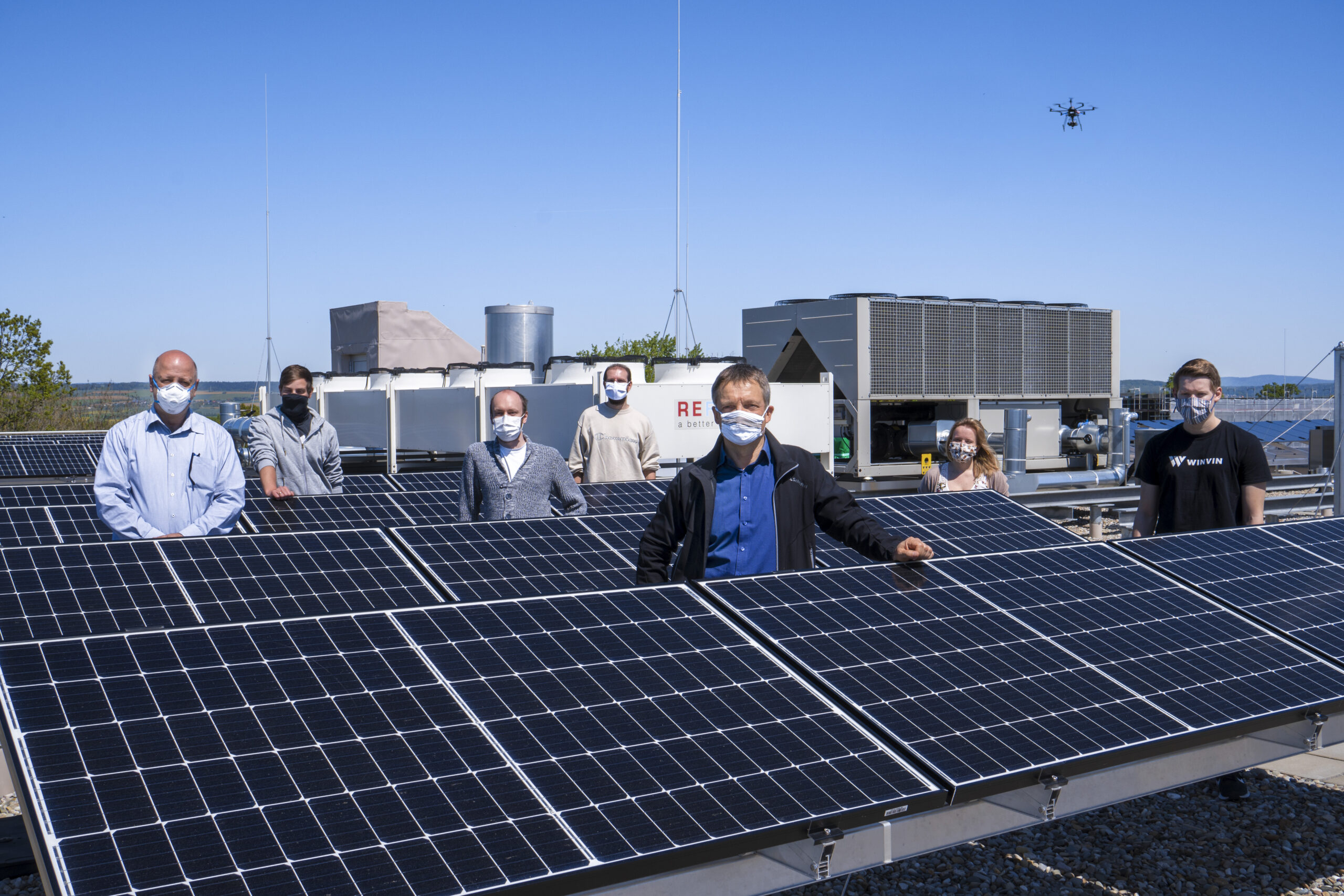 Eine Gruppe von Menschen der Hochschule Coburg steht mit Masken zwischen Solarmodulen auf einem Dach. Im Hintergrund schwebt eine Drohne unter einem klaren blauen Himmel.