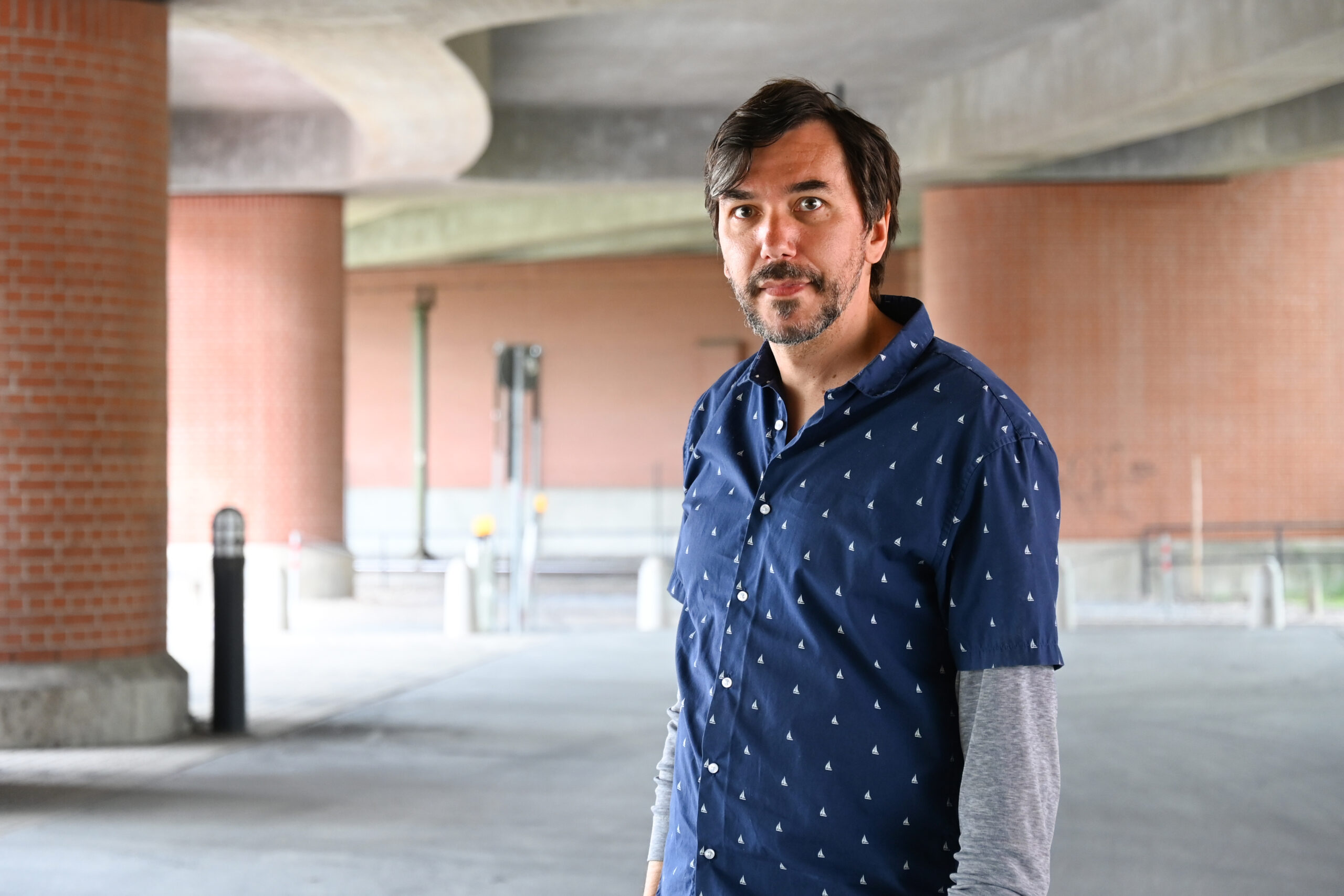 A man with a beard stands in an urban setting under a concrete structure near Hochschule Coburg's brick walls. Wearing a blue patterned shirt over a gray long-sleeve top, he looks toward the camera. The background features architectural elements and muted lighting.