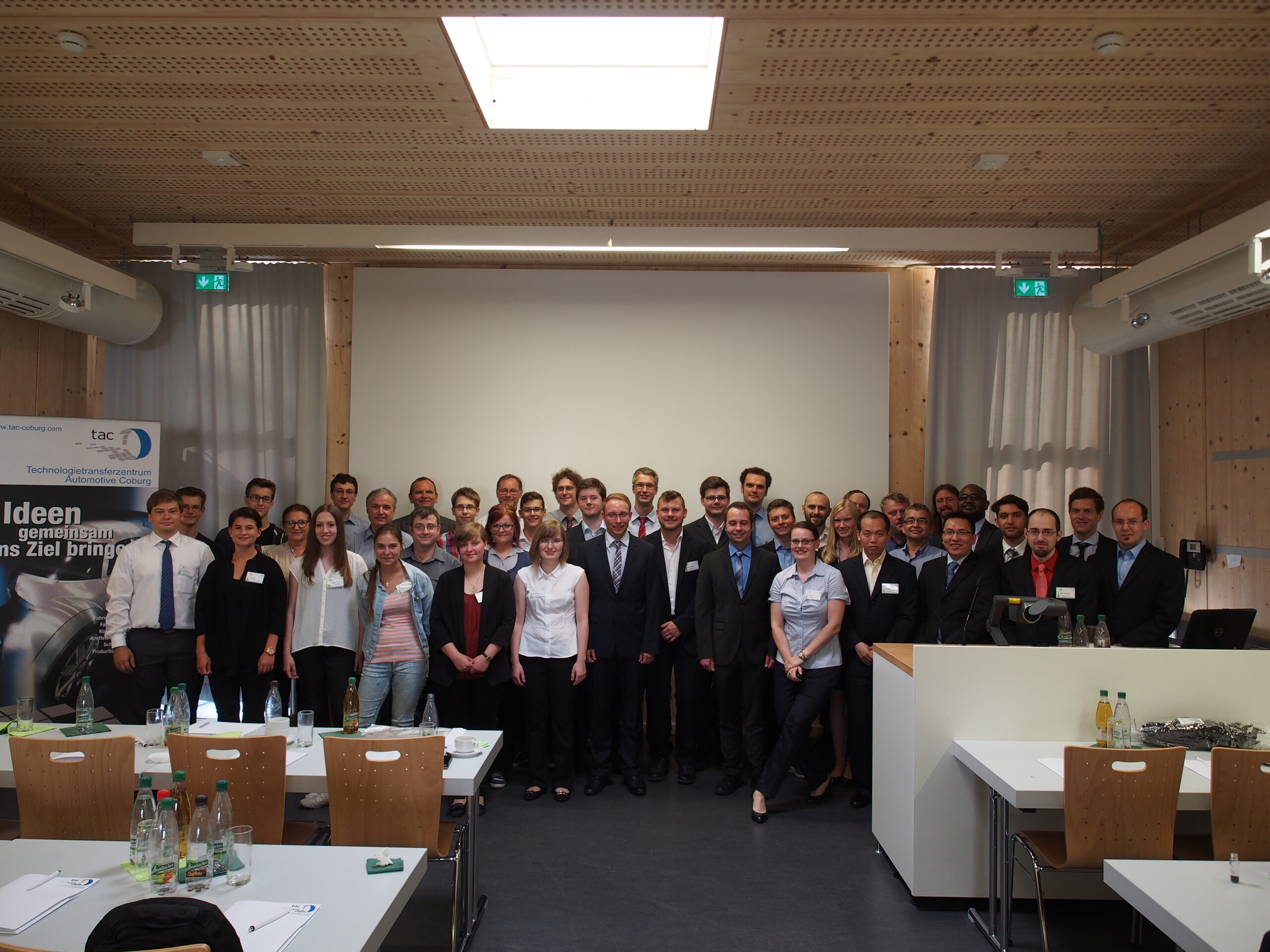 Eine Gruppe von Menschen, einige in Business-Kleidung, andere in Freizeitkleidung, posiert für ein Foto in einem Konferenzraum der Hochschule Coburg. Sie stehen vor einer Präsentationsleinwand, im Vordergrund sind Tische mit Flaschen und Gläsern zu sehen.