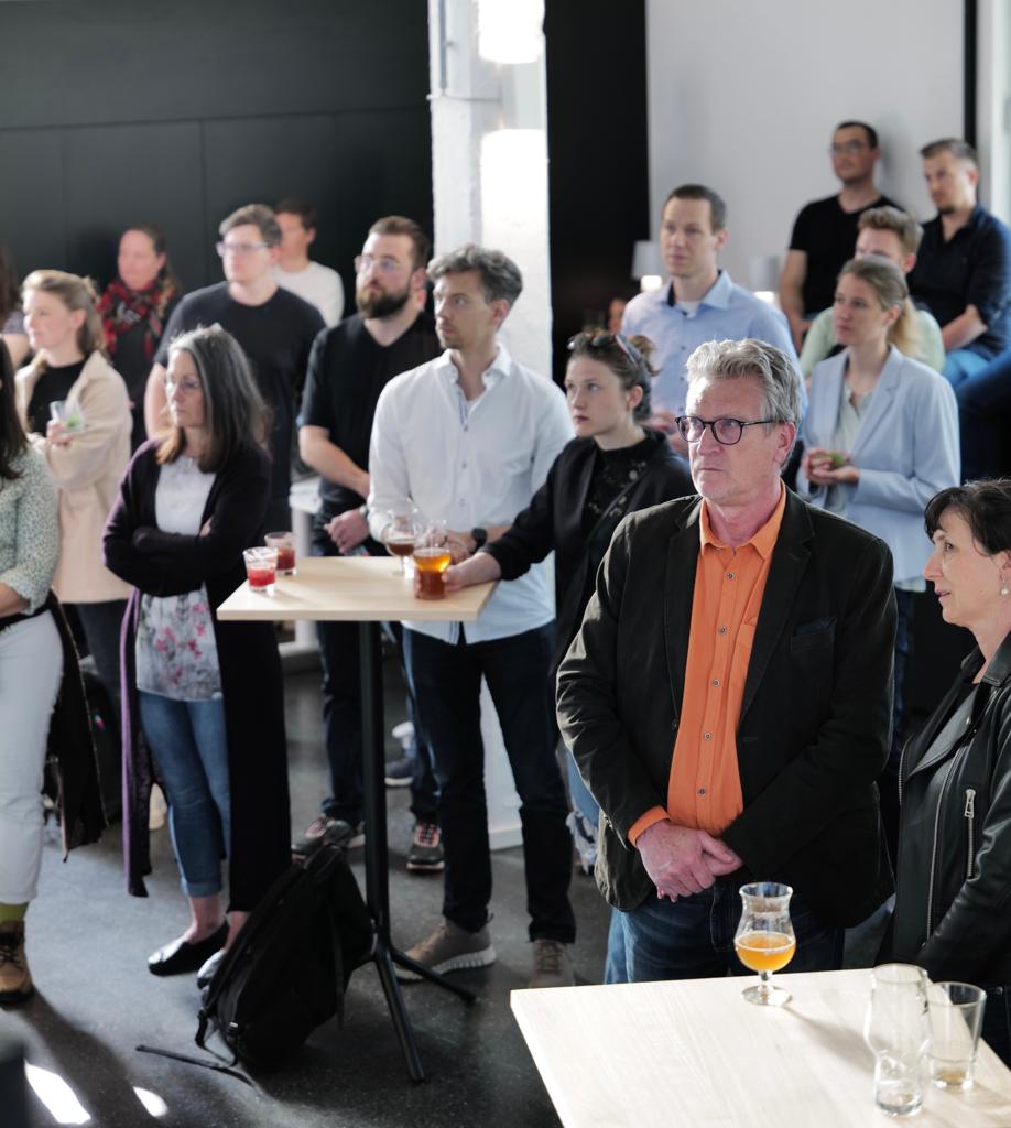 A group of people, possibly from Hochschule Coburg, stand and listen attentively in a modern, well-lit room. Some are seated on high stools, others stand holding drinks. Tables are scattered throughout the space, creating a casual and engaging atmosphere.