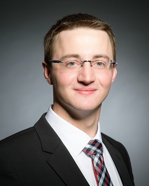 Ein Mann mit kurzen braunen Haaren und Brille lächelt. Er trägt einen schwarzen Anzug, ein weißes Hemd und eine karierte Krawatte und verkörpert damit den professionellen Geist der Hochschule Coburg. Der Hintergrund ist mit einem schlichten Farbverlauf versehen.