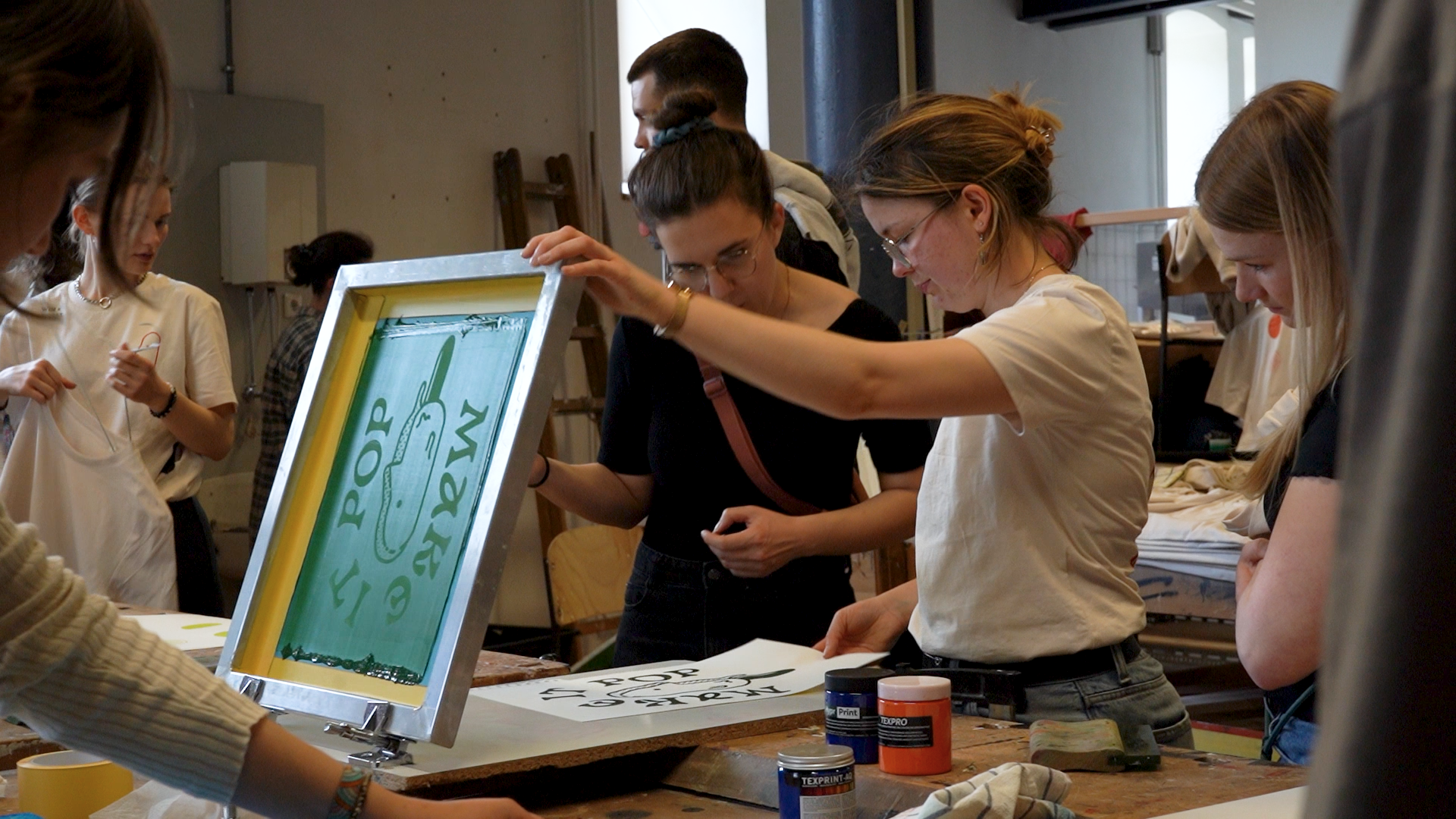 Eine Gruppe von Menschen nimmt an einem Siebdruck-Workshop an der Hochschule Coburg teil. Zwei Frauen im Vordergrund konzentrieren sich auf einen Siebdruck mit den Worten „POP MOVE“. Farbdosen und Werkzeuge sind über den Tisch verteilt, während andere im Hintergrund Kunstwerke herstellen.