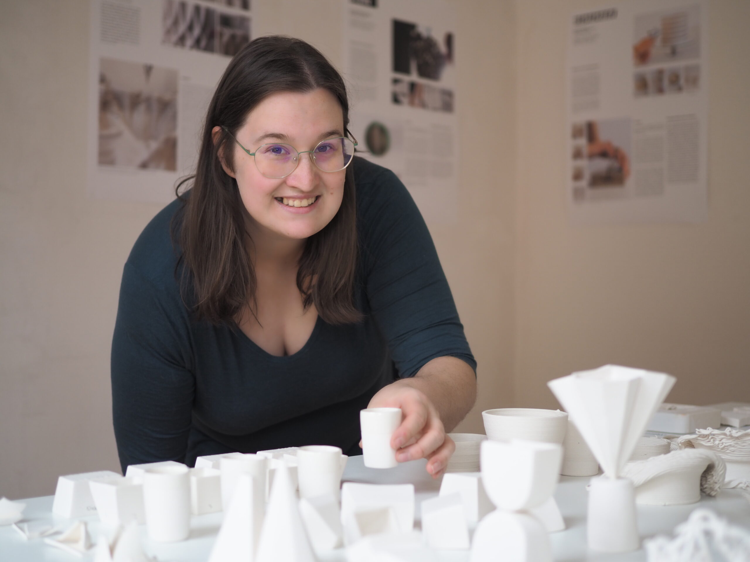 Eine Person mit Brille steht lächelnd neben einem Tisch, der mit geometrischen weißen Tonmodellen bedeckt ist, an der Hochschule Coburg. Im Hintergrund hängen gedruckte Informationsblätter an der Wand.