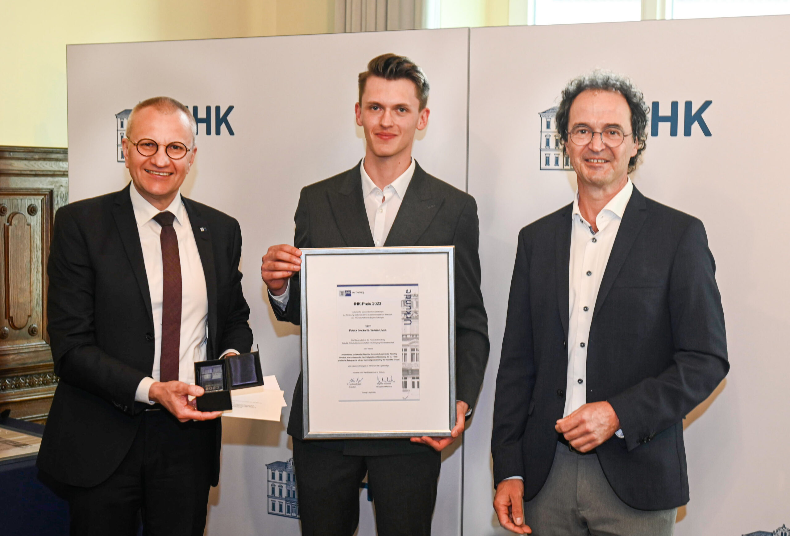 Three men stand together, smiling. The middle man holds a large framed certificate, while the man on the left holds a small box with a medal. They are in a formal setting with an IHK logo and a Hochschule Coburg banner in the background.