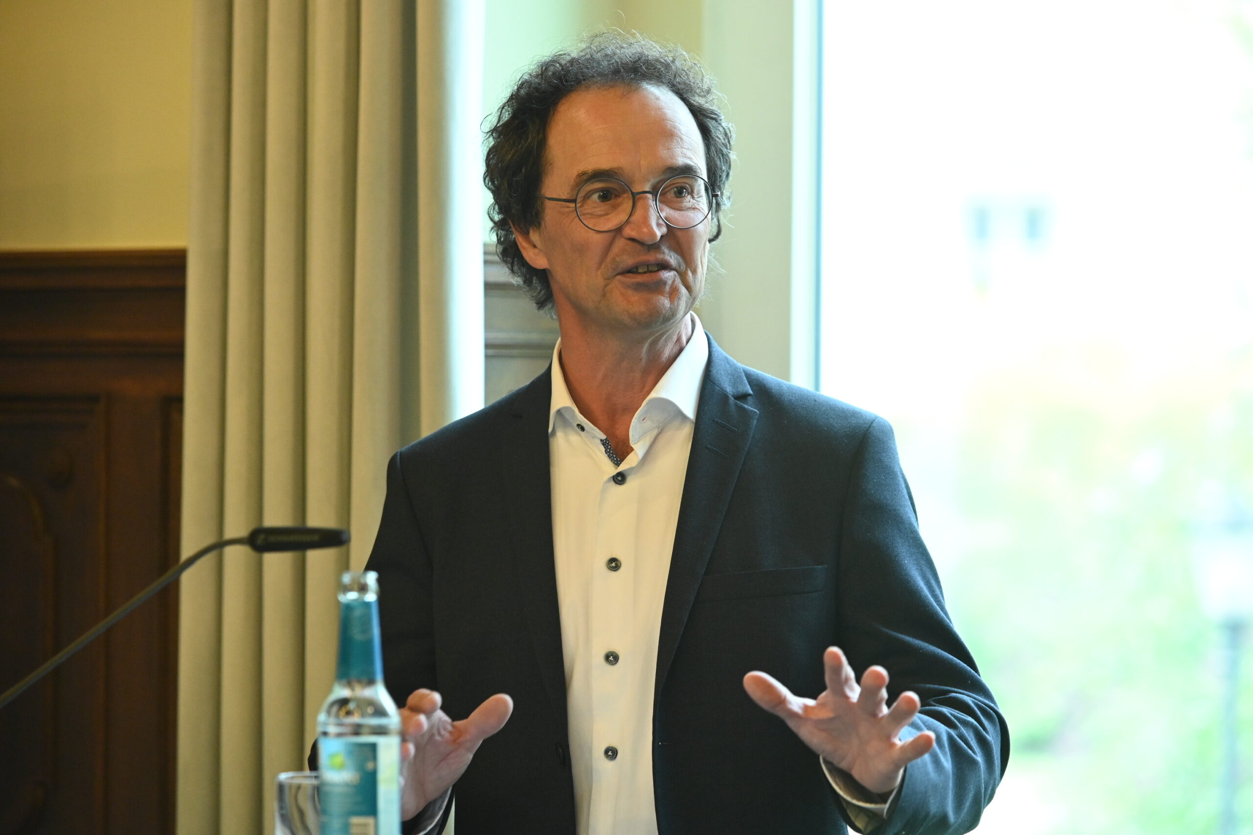 A man with glasses and curly hair is speaking in front of a window at Hochschule Coburg. He wears a dark suit jacket and a white shirt, holding his hands up. A bottle and a microphone are on the table beside him.