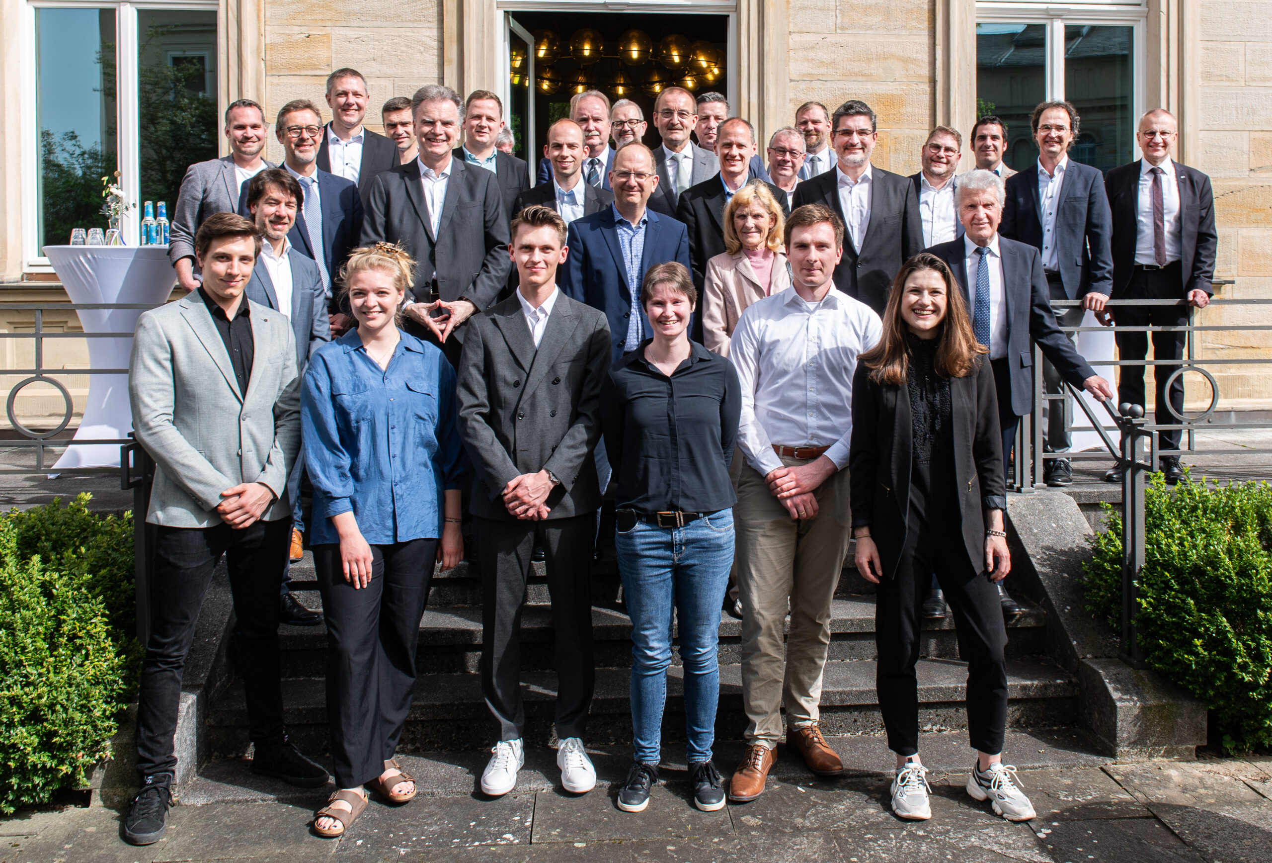 Eine Gruppe von Männern und Frauen hat sich auf den Außentreppen der Hochschule Coburg versammelt. Sie sind in Business- und Freizeitkleidung gekleidet. Sie stehen vor einem Gebäude mit großen Fenstern und lächeln in die Kamera. In der Nähe gibt es kleine Büsche und einen Tisch.