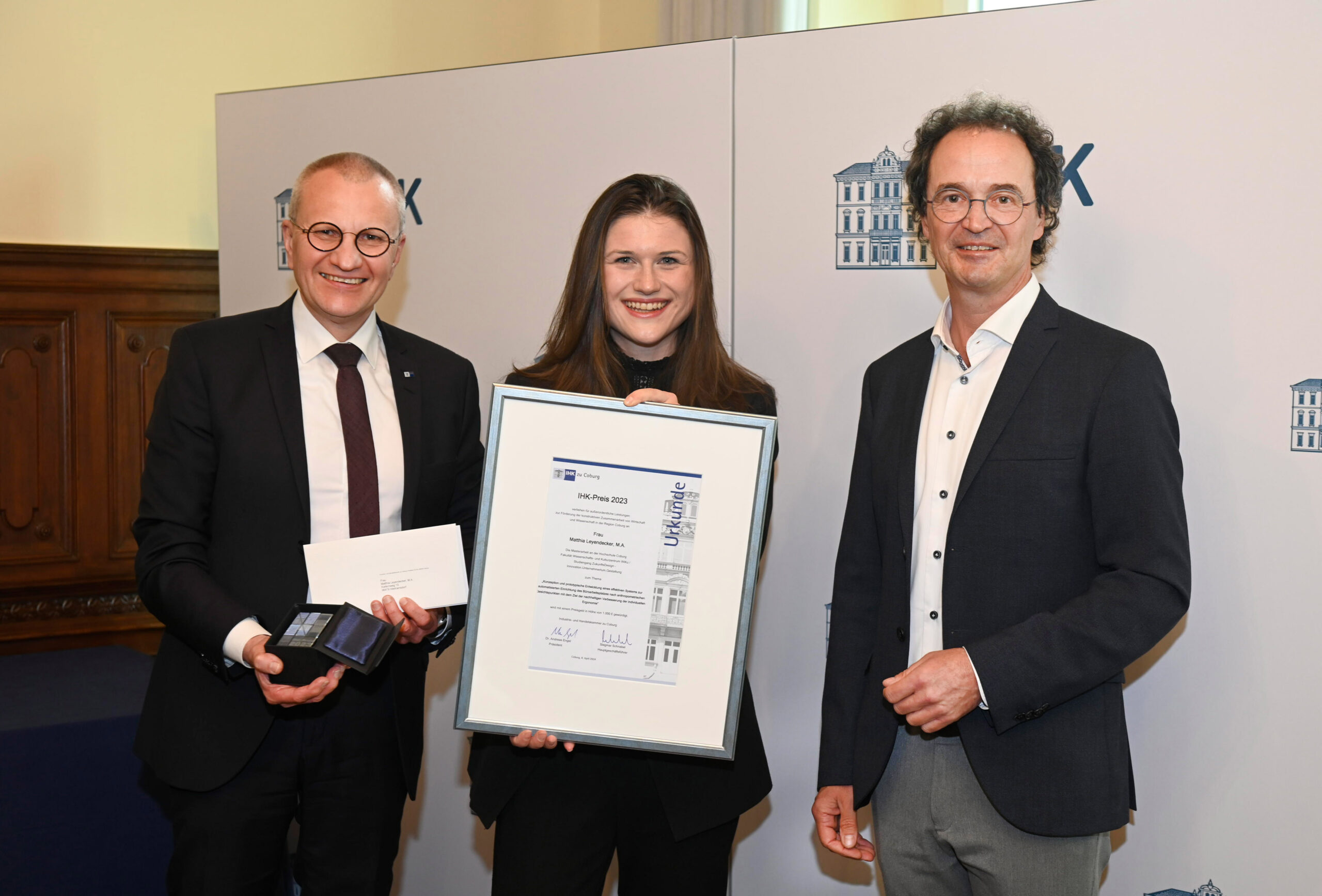 Three people stand together smiling. The person in the middle holds a framed certificate from Hochschule Coburg. The person on the left carries an envelope and a small box. In the background, a white banner with blue graphics of a building completes the celebratory scene.