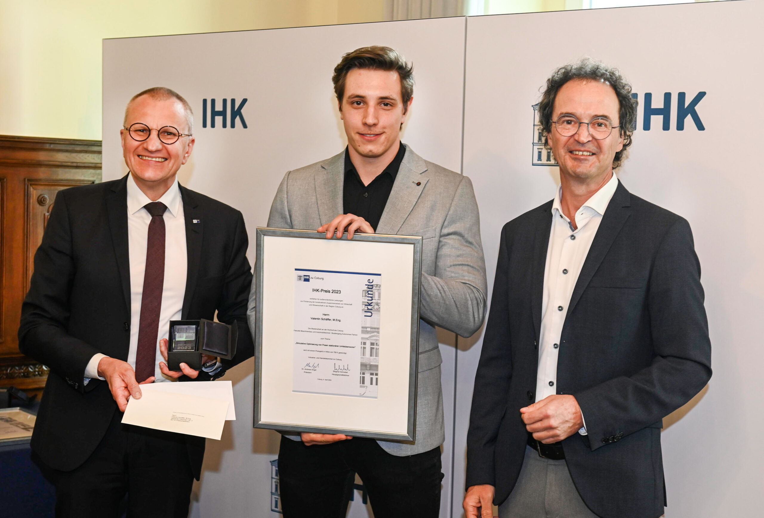 Three men stand facing the camera. The man in the middle holds a framed certificate, while the man on the left presents an open box. They are positioned in front of a backdrop featuring "IHK" and "Hochschule Coburg.