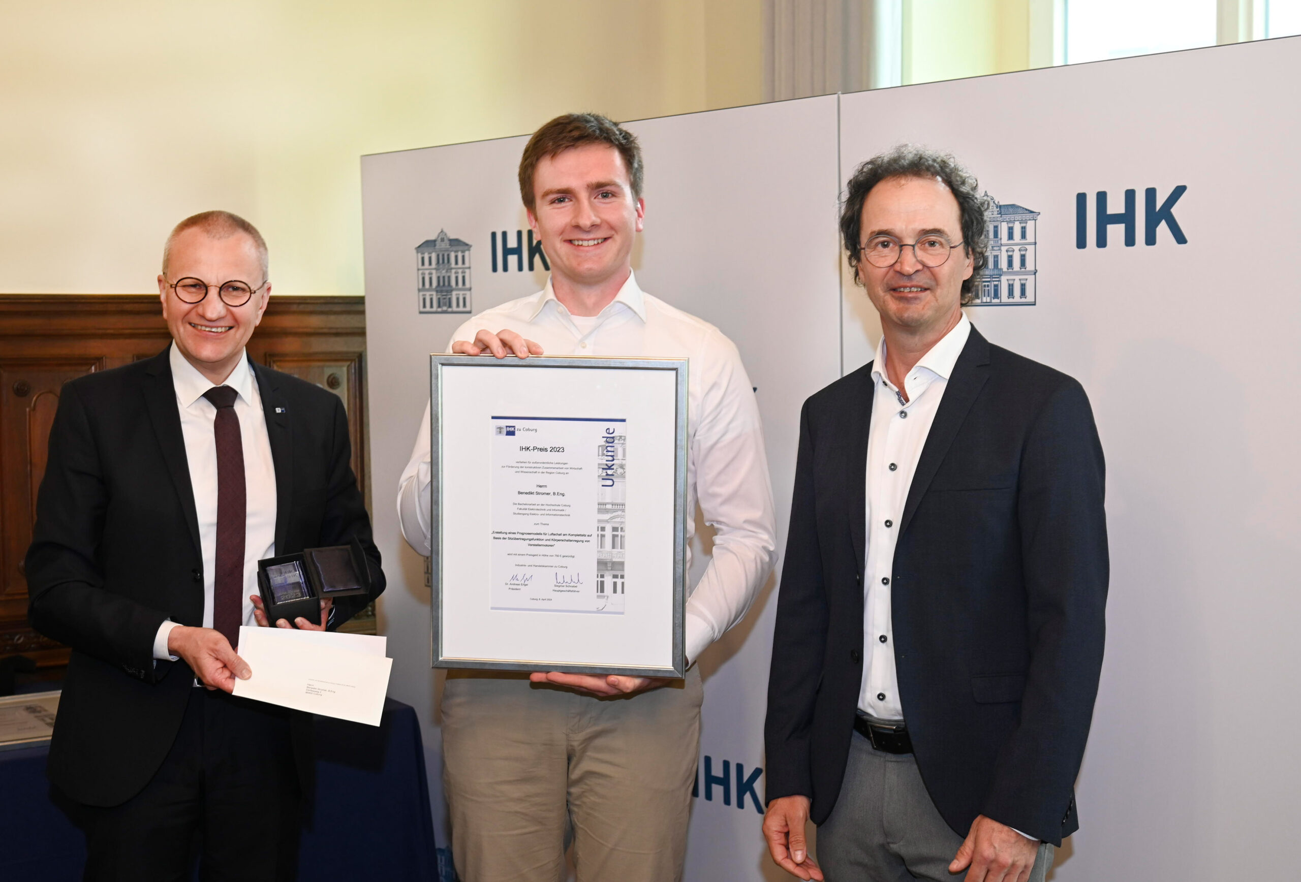 Three men stand together, smiling. The man in the middle holds a framed certificate, marking their achievement with Hochschule Coburg. Another man on the left holds a small plaque and an envelope. A backdrop with the IHK logo is visible in the background.