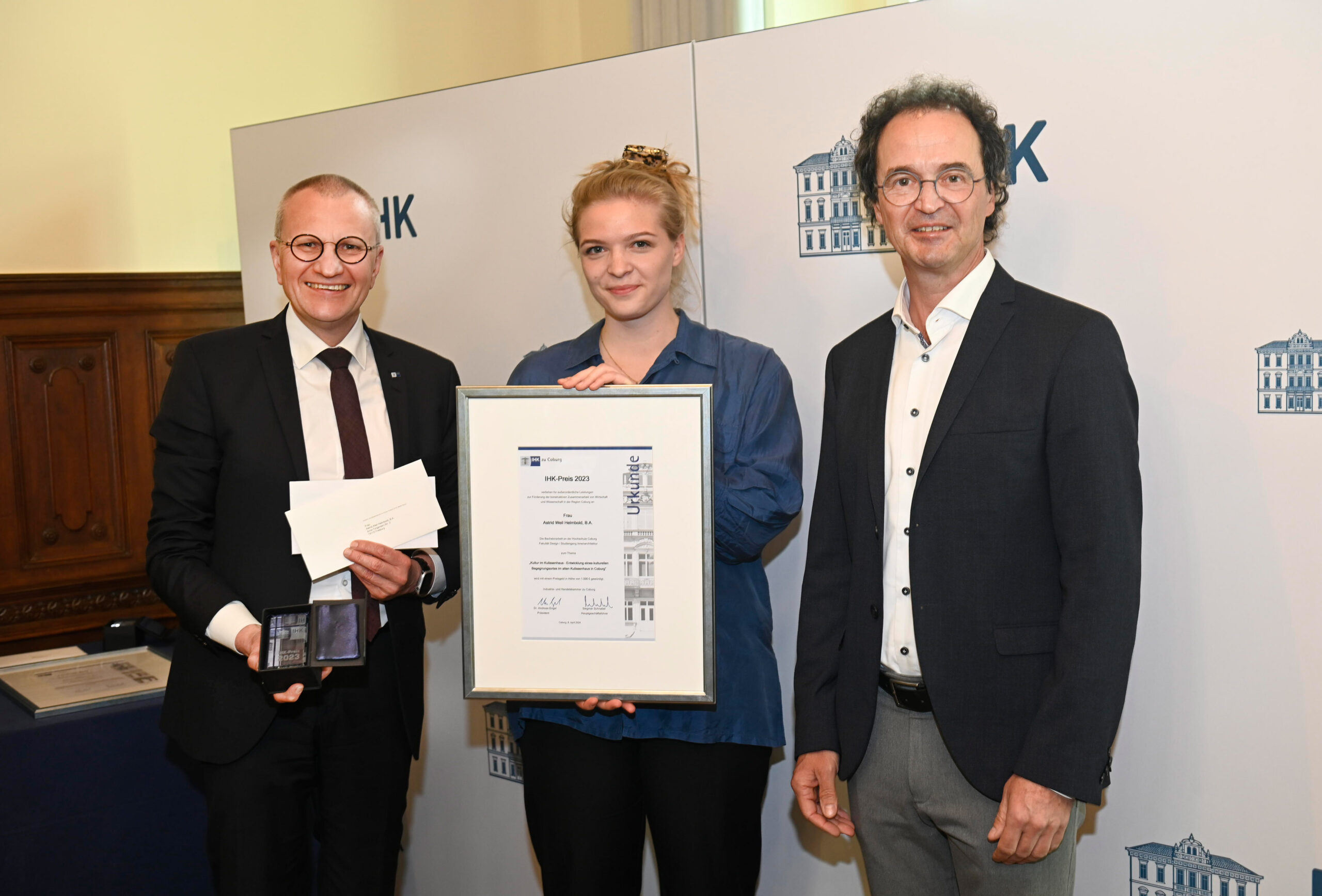 Three people stand indoors, smiling at the camera. The person in the middle holds a framed certificate from Hochschule Coburg. The person on the left holds cards or papers. They are standing in front of a backdrop with a building design, capturing a memorable academic moment.