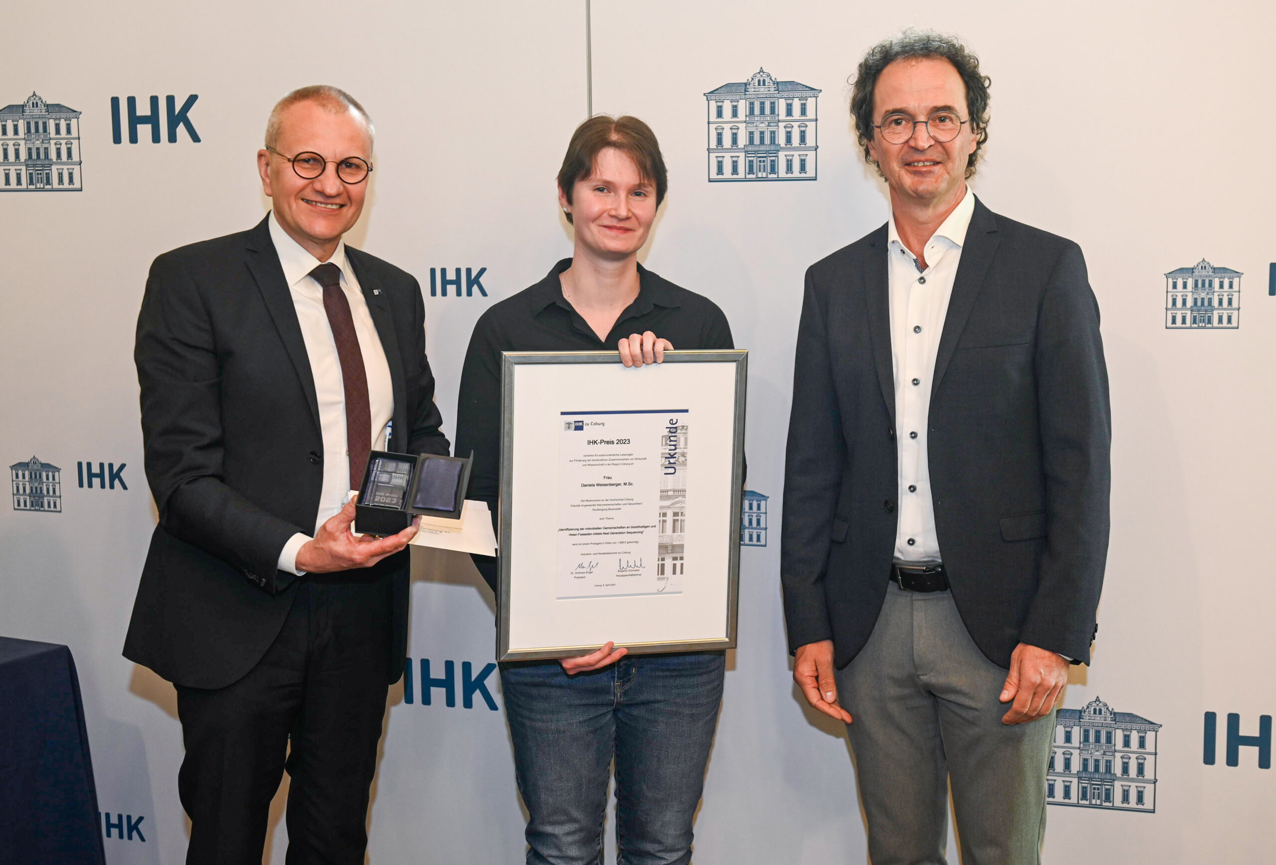 Three people are standing in front of a backdrop featuring "IHK" and building illustrations. The person in the middle, representing Hochschule Coburg, holds a framed certificate while another shows an open box with a medal. They are all dressed in business attire.