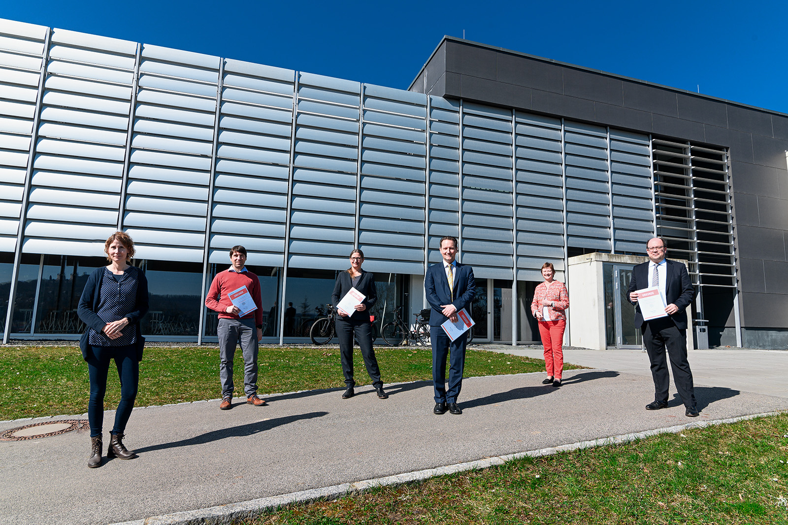 Eine Gruppe von sechs Personen steht in einer Reihe vor dem modernen Gebäude der Hochschule Coburg mit großen Fenstern und glatten Metallpaneelen. Sie halten Ordner in den Händen und wahren unter dem klaren blauen Himmel Abstand zueinander.