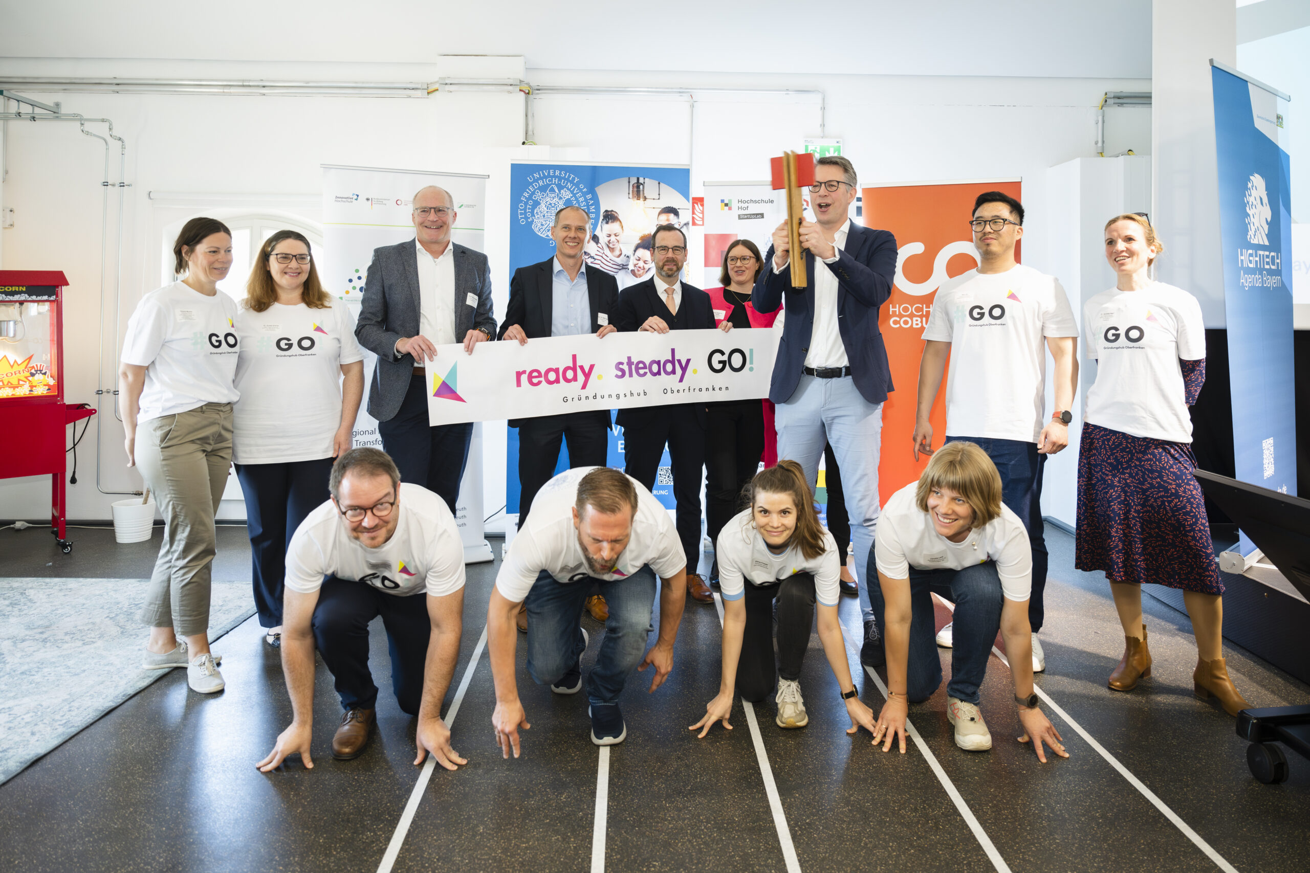 A group of people at Hochschule Coburg participate in a playful indoor activity. Some are crouched in a starting position on a mocked-up track, while others hold a "ready. steady. GO!" banner and smile amidst colorful decorations in the background.