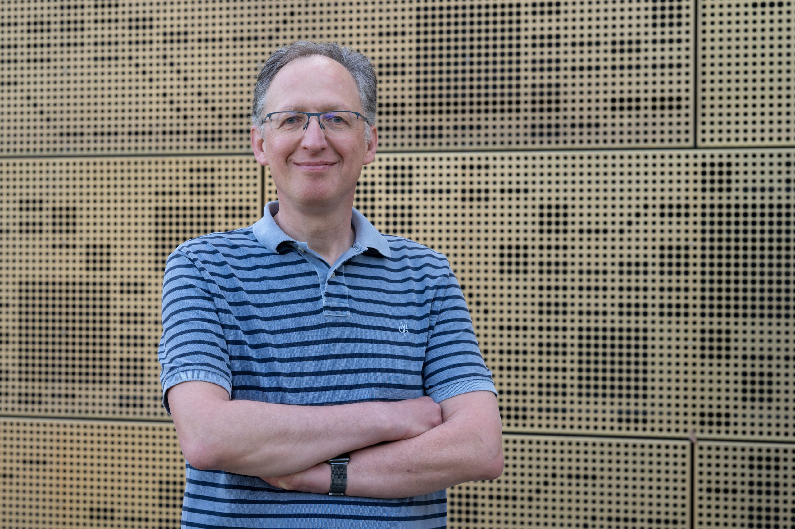 Eine Person mit Brille und gestreiftem Poloshirt, die den selbstbewussten Geist der Hochschule Coburg verkörpert, steht lächelnd und mit verschränkten Armen vor einer strukturierten Wand.