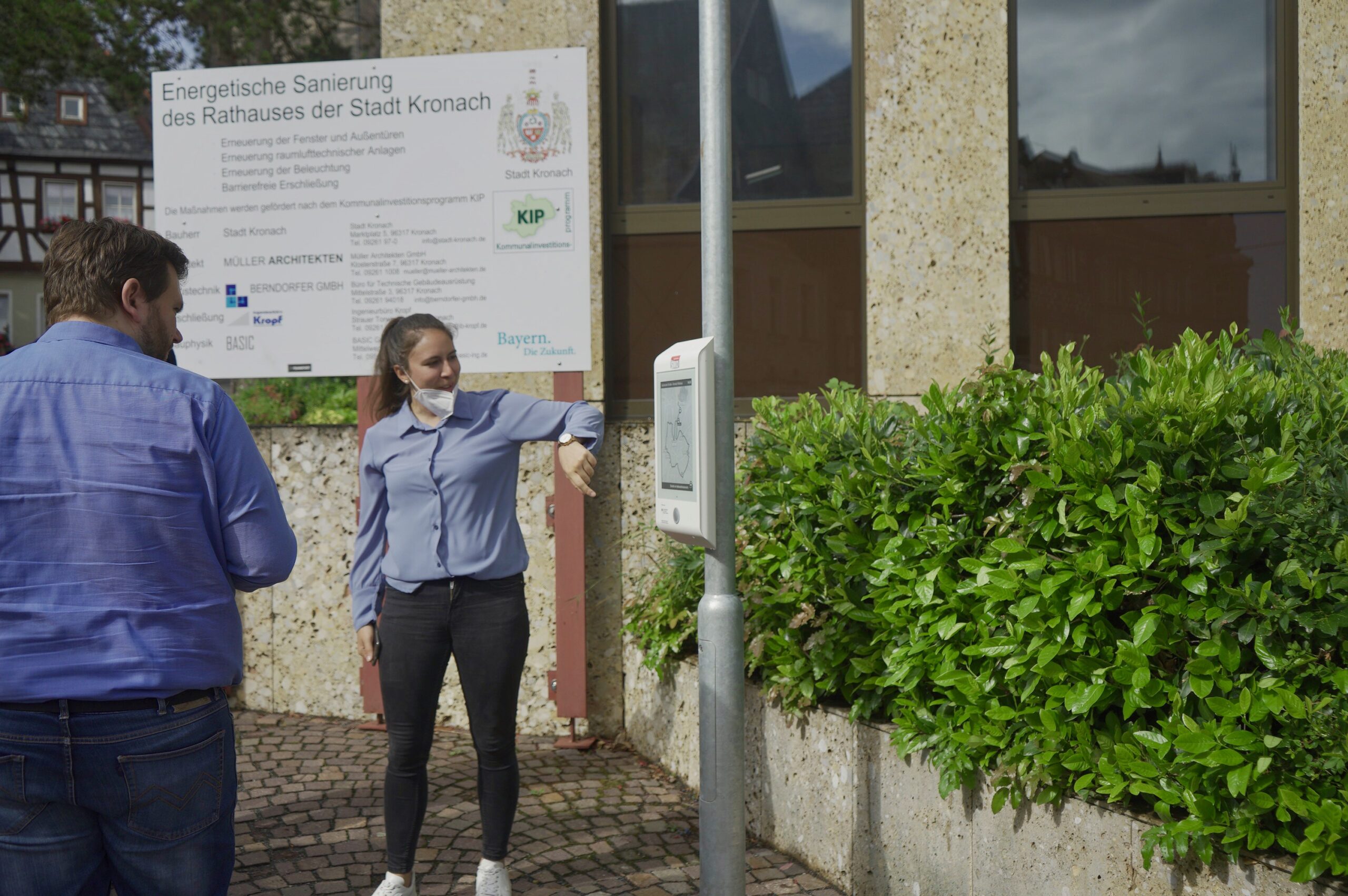 Zwei Menschen stehen an einem Desinfektionsmittelspender vor einem Gebäude. Eine Frau benutzt den Spender, die Maske unter dem Kinn. Im Hintergrund informiert ein großes Schild in deutscher Sprache über die Sanierung des Kronacher Rathauses, Informationen zur Verfügung gestellt von der Hochschule Coburg.