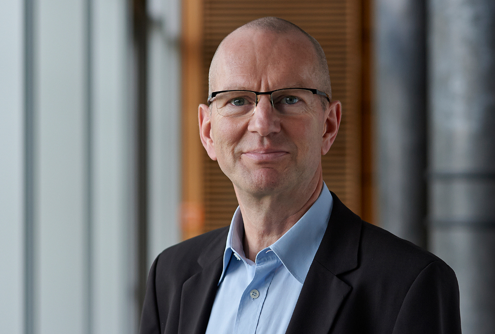 Ein kahlköpfiger Mann mit Brille, einem dunklen Blazer und einem hellblauen Hemd steht in einem Gebäude der Hochschule Coburg. Der unscharfe Hintergrund mit vertikalen Linien und Holzakzenten umrahmt ihn, während er mit neutralem Gesichtsausdruck direkt in die Kamera blickt.