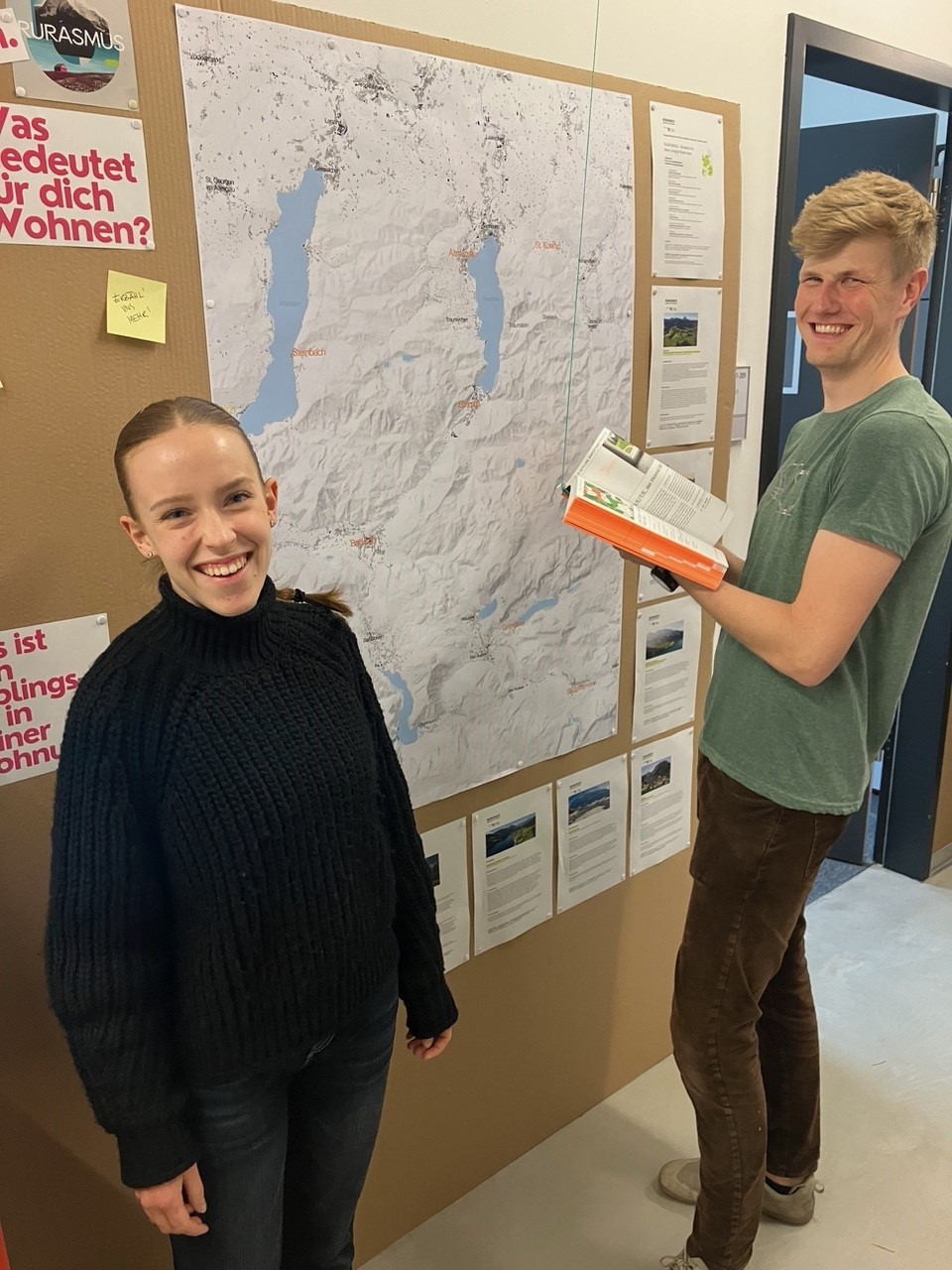 Two smiling people stand in front of a large map on a bulletin board at Hochschule Coburg. One holds a book and points to the map, while various papers and notes are pinned around it. Bright indoor lighting highlights their engaging interaction.