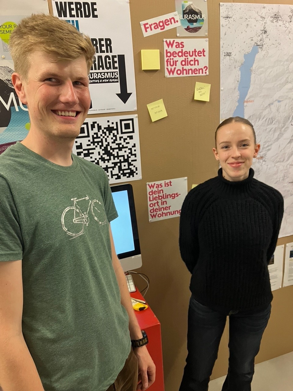 Two people stand smiling by a wall adorned with Hochschule Coburg posters, sticky notes, and a map. One wears a green shirt with a bicycle graphic; the other sports a black sweater. A computer monitor is visible between them.