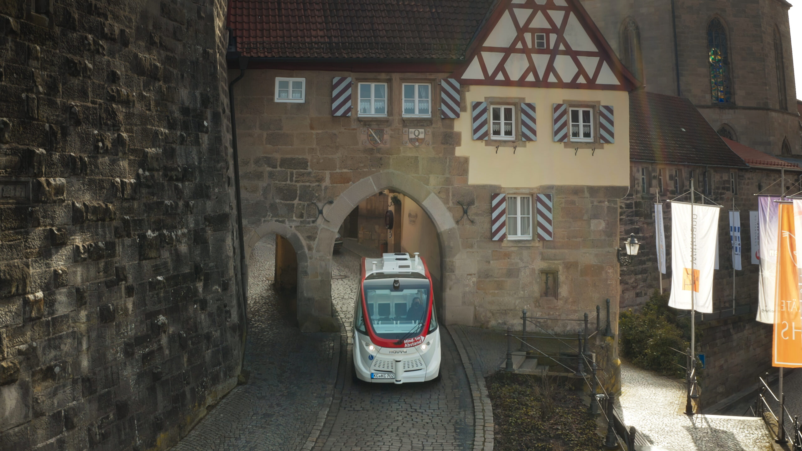 Ein autonomes Fahrzeug gleitet durch einen mittelalterlichen Steinbogen in einer historischen Stadt. Darüber trägt ein traditionelles Fachwerkgebäude mit kleinen Fenstern und roten Fensterläden das Emblem der Hochschule Coburg, alles unter einem klaren Himmel.