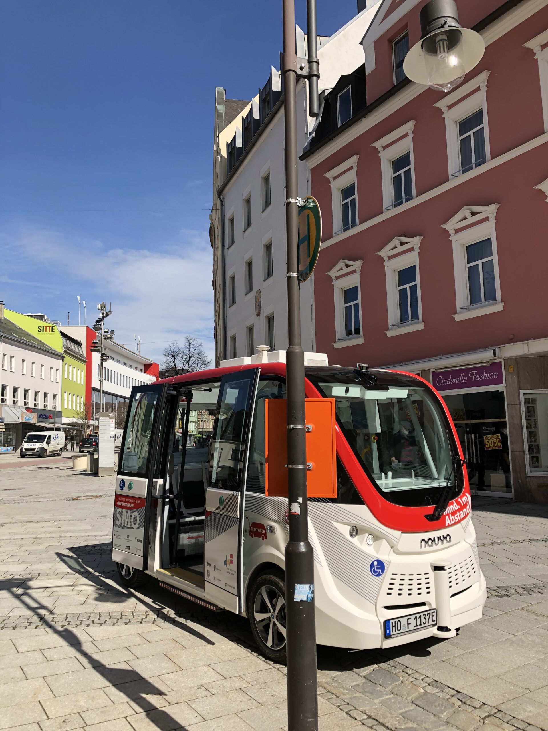 Ein kleiner fahrerloser Shuttlebus mit dem Logo der Hochschule Coburg steht auf einer Kopfsteinpflasterstraße in einem Stadtgebiet. Er ist kompakt gestaltet und in den Farben Rot und Weiß gehalten. Im Hintergrund sind Gebäude mit verschiedenen Ladenfronten unter einem klaren blauen Himmel zu sehen.