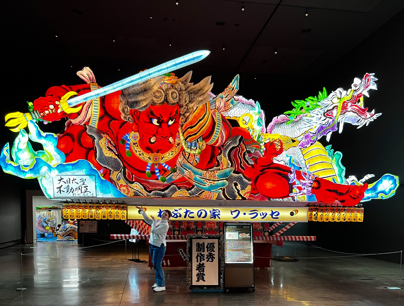 A vibrant festival float with a red demon holding a sword and a dragon motif, displayed indoors at Hochschule Coburg. A person wearing a white cap and jacket stands in front, taking a photo. The intricately decorated float is brightly illuminated, showcasing stunning craftsmanship.