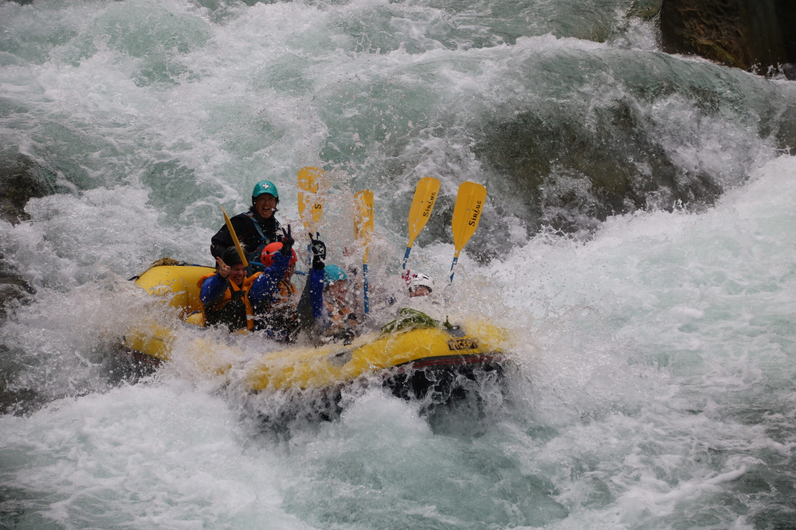 A group of people from Hochschule Coburg, wearing helmets and life jackets, navigates a yellow inflatable raft through turbulent white-water rapids, raising their paddles in synchronized motion amid splashing waves.