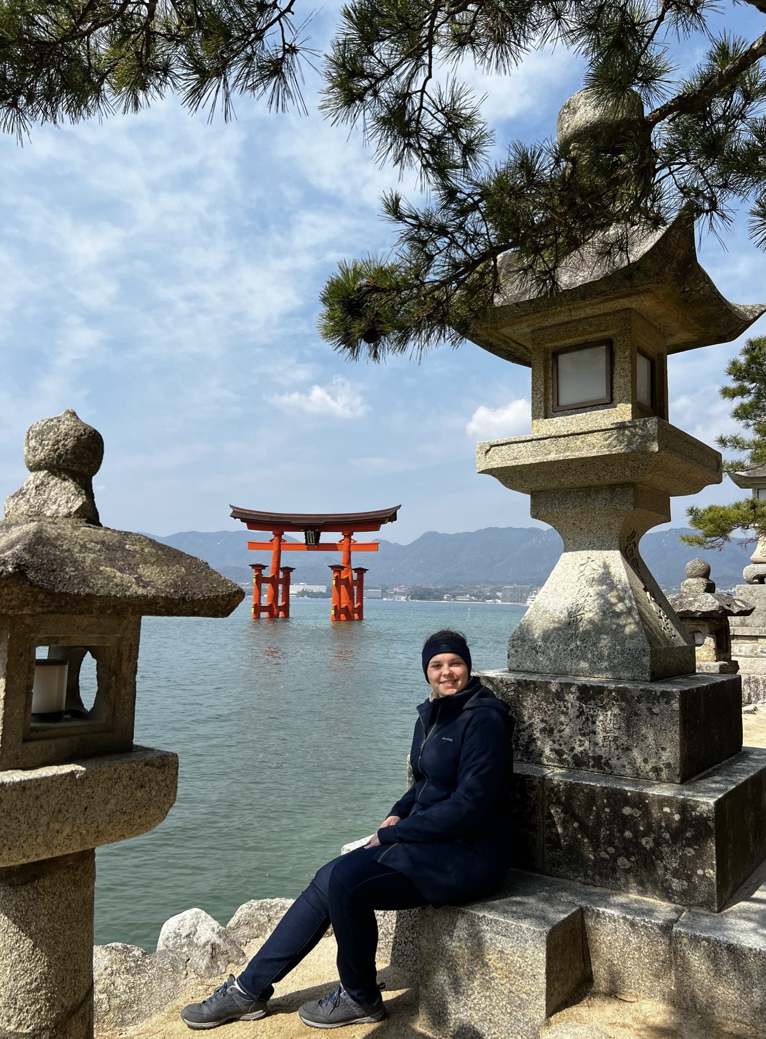 A person in a dark jacket and hat sits on a stone wall by the waterfront, reminiscent of the peaceful ambiance one might find near Hochschule Coburg. In the background, a large red torii gate stands in the water, surrounded by scenic hills, with stone lanterns and pine branches framing the scene.