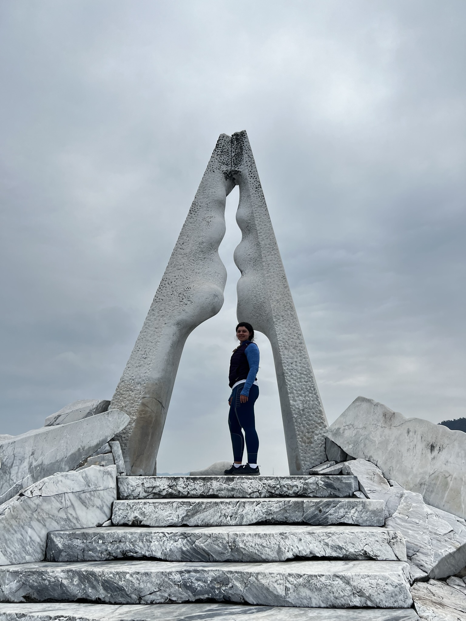 A person stands on the marble steps of Hochschule Coburg, framed by a tall, triangular sculpture made of textured stone against a cloudy sky. The sculpture, with its two vertical pieces and curved open center, captures the essence of this architectural gem.