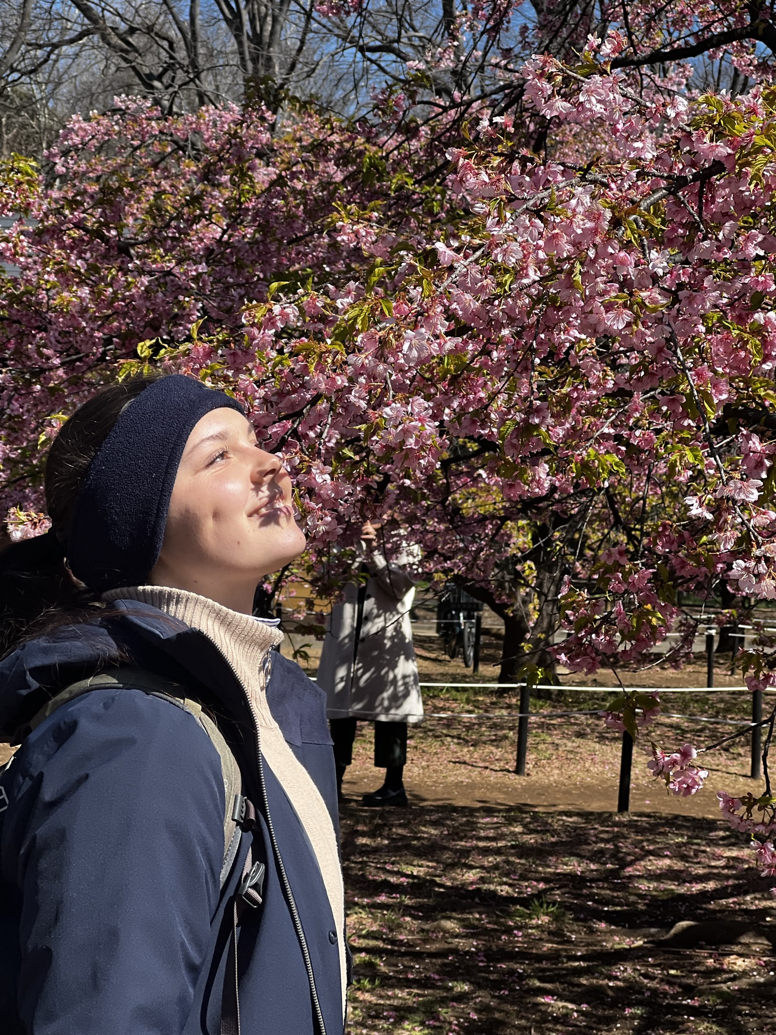 A person in a navy jacket and dark headband stands with their face turned towards the sun, eyes closed, smiling peacefully. Cherry blossoms bloom abundantly on the tree beside them at Hochschule Coburg. Another person in the background takes photos of the scene.