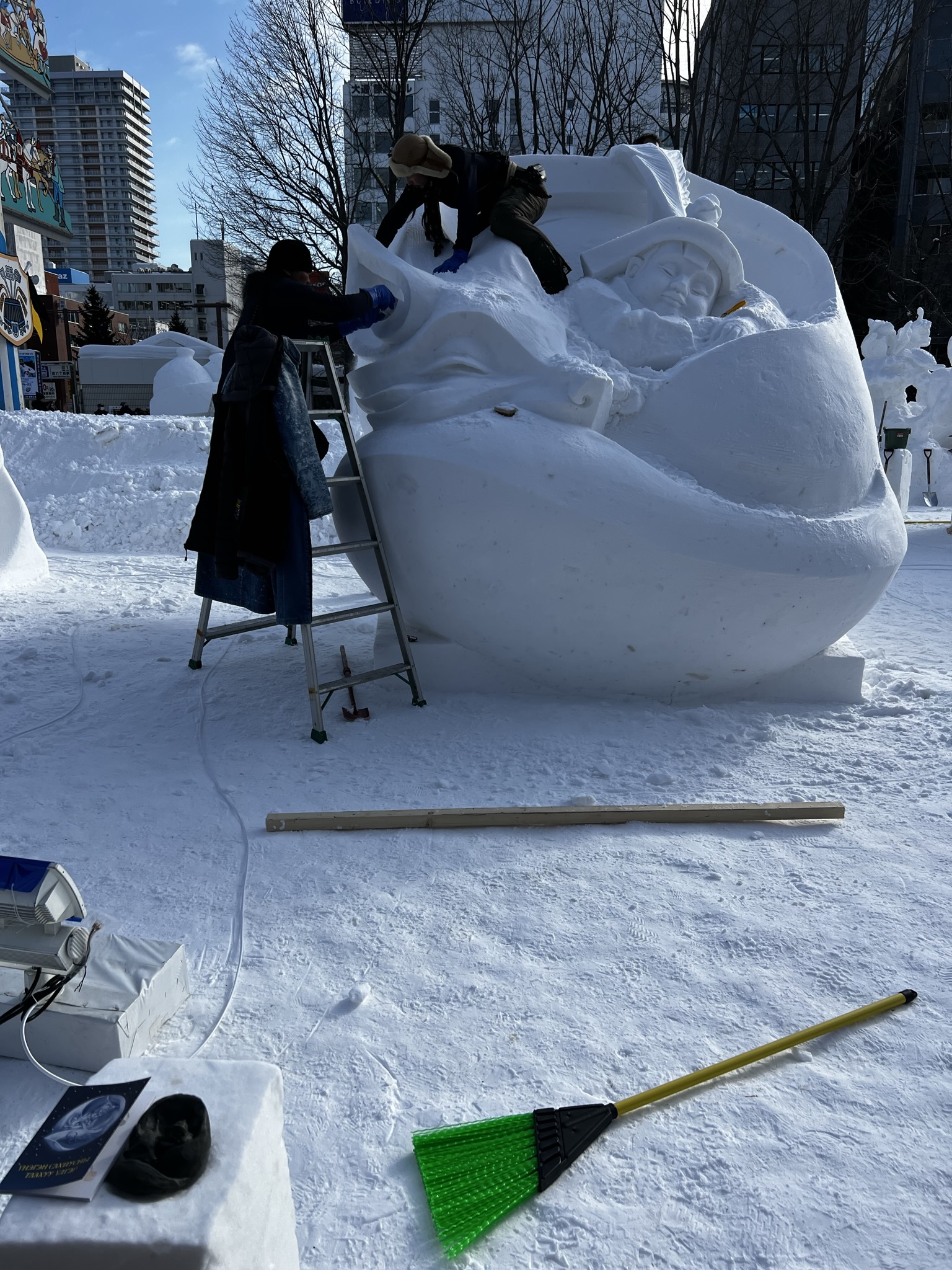 Two people work on a large snow sculpture near Hochschule Coburg, featuring faces and intricate designs. One stands on a ladder, while the other sits atop the creation. Nearby are a green broom, equipment, and a book resting on the snowy ground.