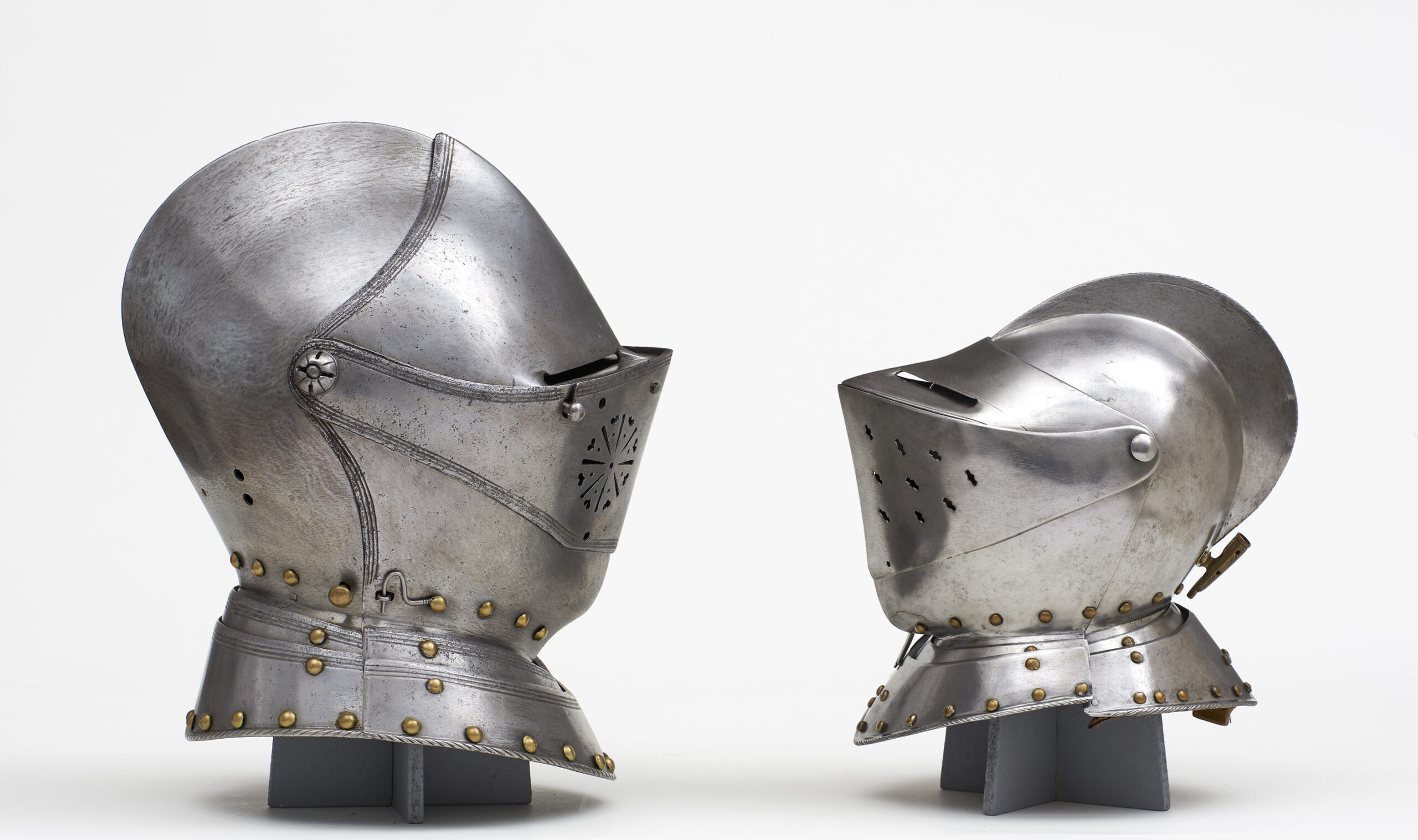 Two medieval steel helmets facing each other, displayed on stands, evoke a sense of history at Hochschule Coburg. The helmets, adorned with metal studs and visors, showcase intricate craftsmanship against a plain white background.