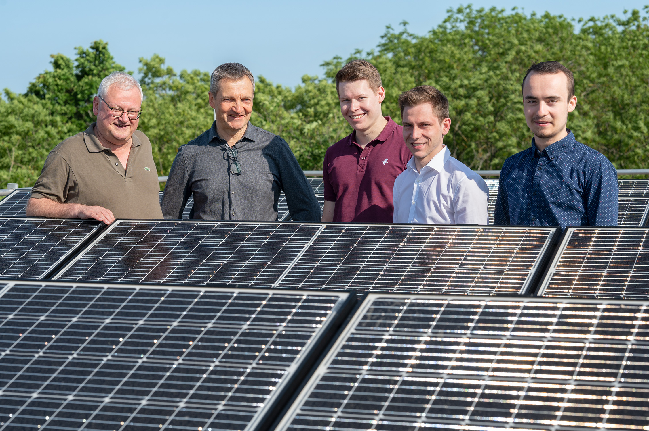 Fünf Männer der Hochschule Coburg stehen gemeinsam lächelnd hinter Solarmodulen inmitten des üppigen Grüns und unter einem strahlend blauen Himmel.