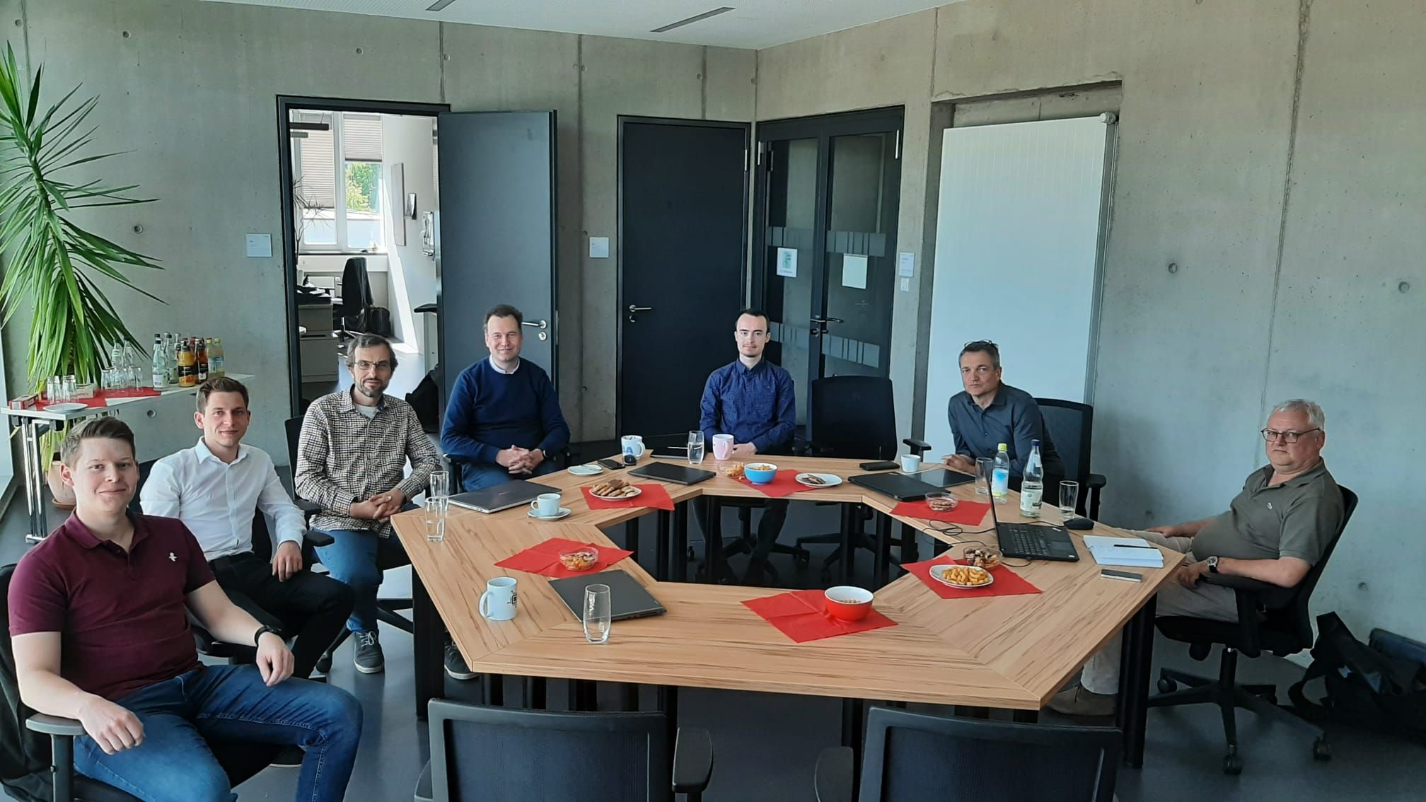 Sieben Personen sitzen in einem modernen Konferenzraum der Hochschule Coburg um einen großen, L-förmigen Tisch, der mit Laptops und Unterlagen ausgestattet ist. Am Tisch stehen Snacks und Getränke. In der Ecke ist eine Topfpflanze zu sehen, und die hellgrauen Wände des Raums fördern eine Atmosphäre innovativer Zusammenarbeit.