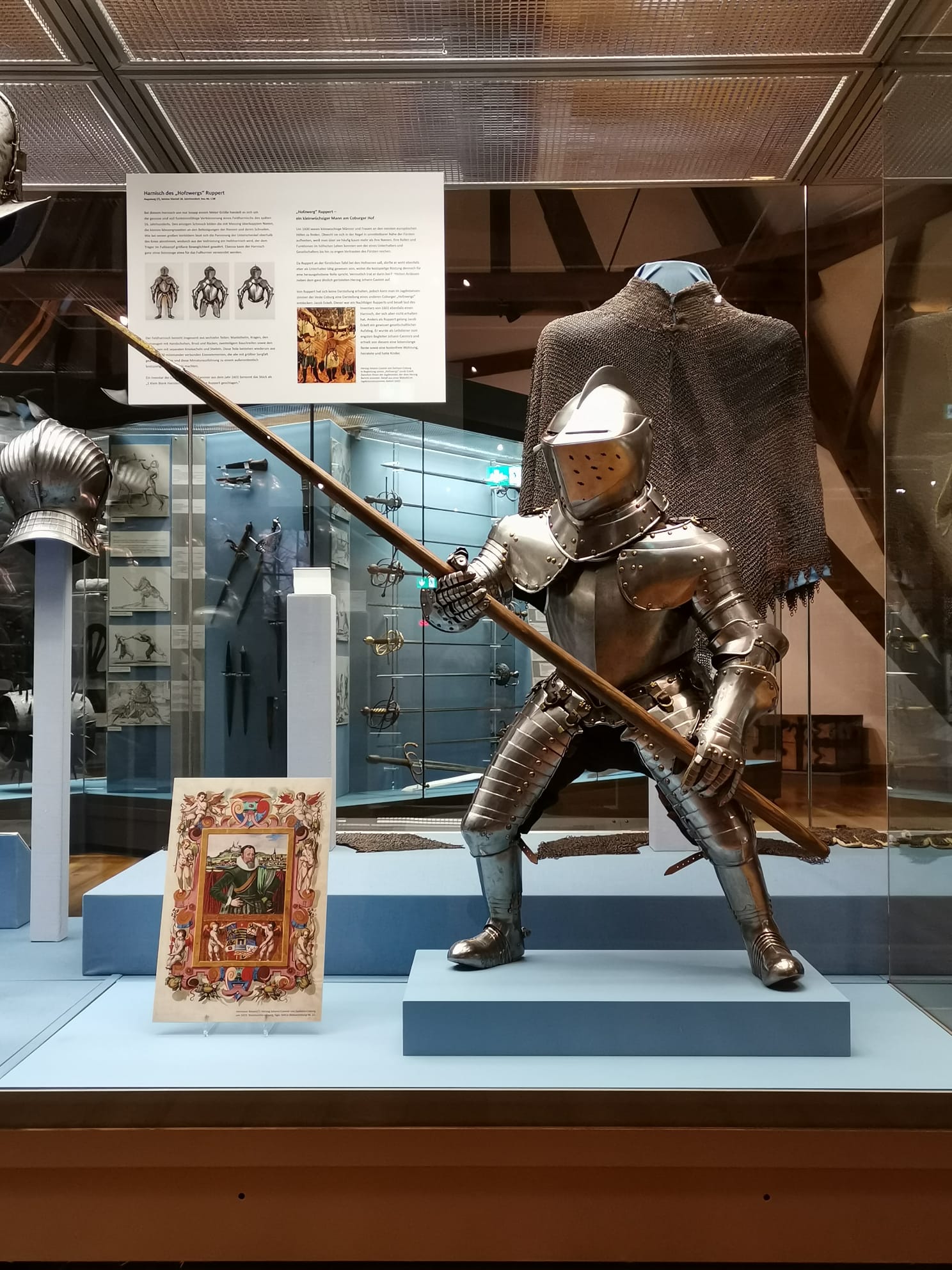 A museum exhibit at Hochschule Coburg displays a suit of medieval armor, complete with a helmet and lance, in a striking pose. Behind the armor, informational plaques and more artifacts are visible, while a colorful, illuminated manuscript page is displayed in front.