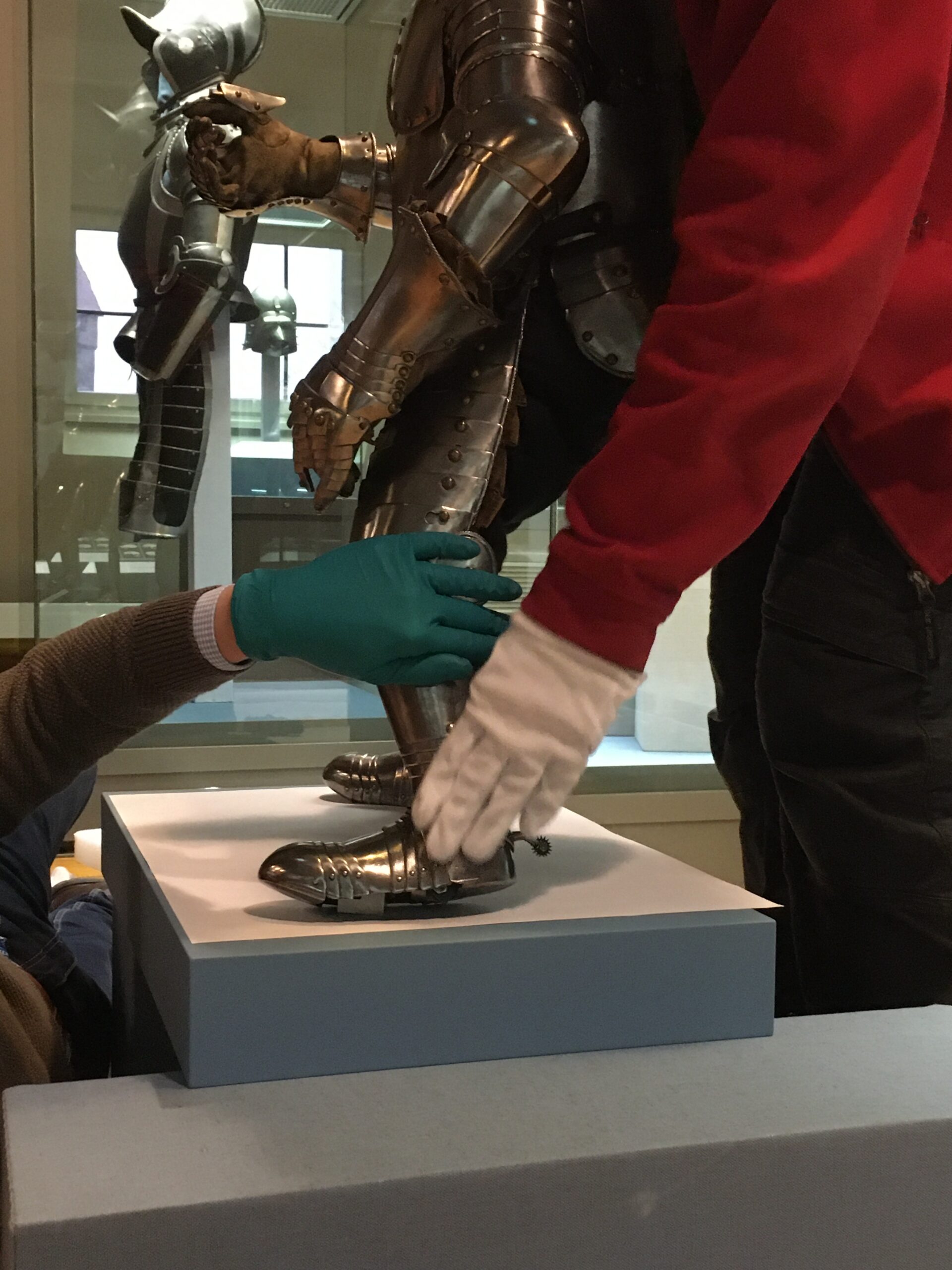 Two people wearing gloves, possibly from Hochschule Coburg, adjust a suit of medieval armor on a pedestal. One wears green gloves and the other white. They carefully position the armor's leg and shoe, with a larger armored figure looming in the background.